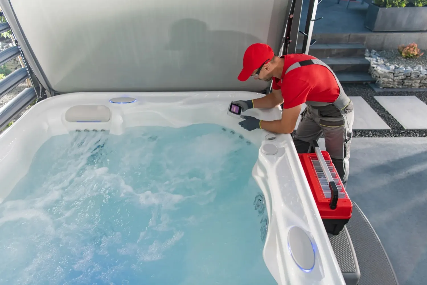 A gloved technician in a red shirt, red baseball cap, and overalls is using a handheld device to service a hot tub with bubbling water on an outdoor patio.