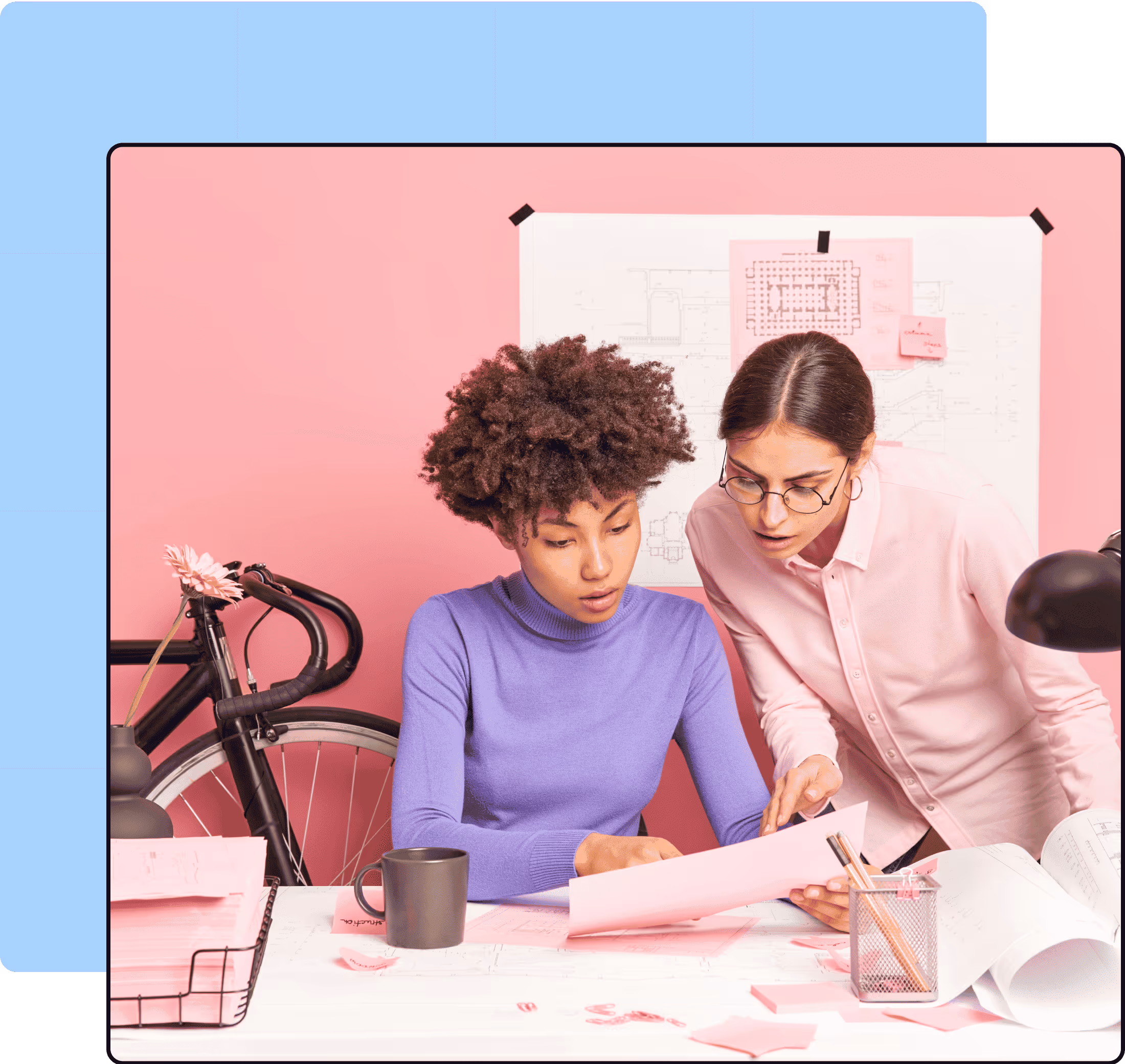 Two women collaborating on architectural or design plans at a desk with pink and purple decor, surrounded by blueprints, a bicycle, and office supplies.