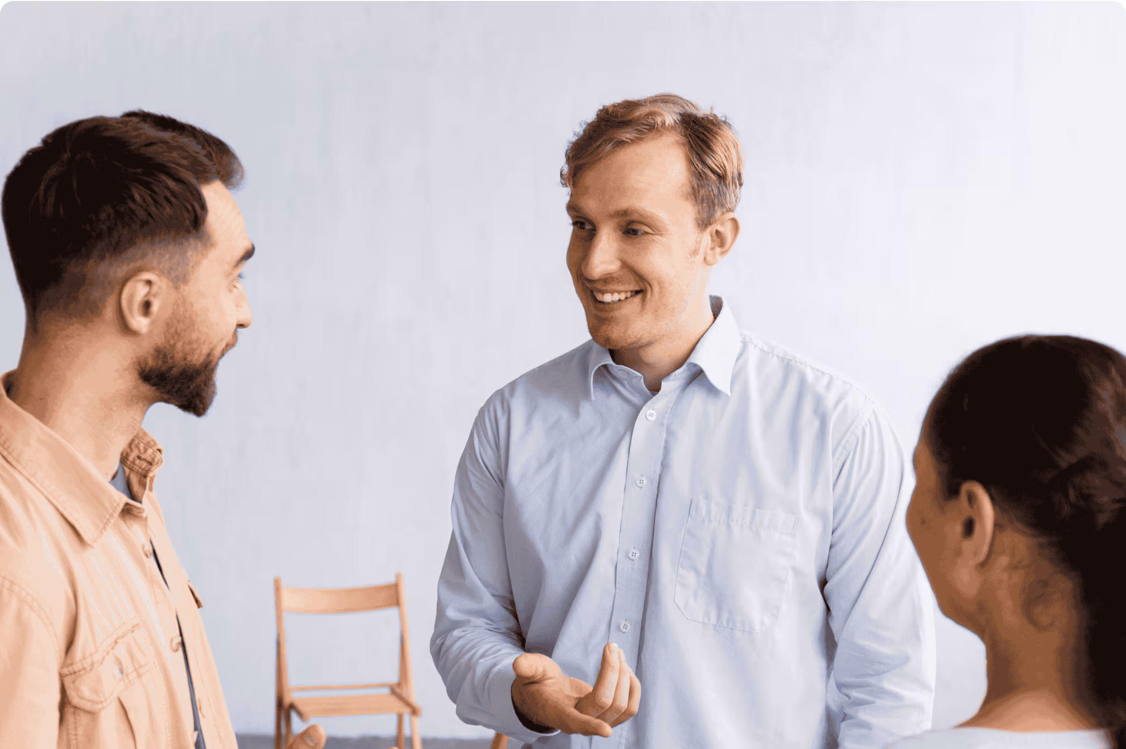 Smiling man in a light blue shirt talking with two people in a casual indoor setting, suggesting a friendly team or client conversation.