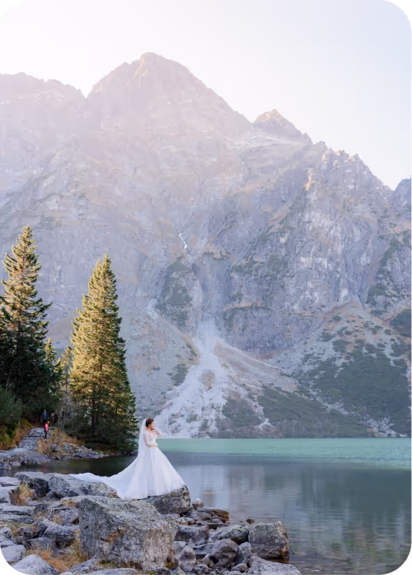Bride in a white wedding dress standing on rocks by a mountain lake with tall peaks in the background