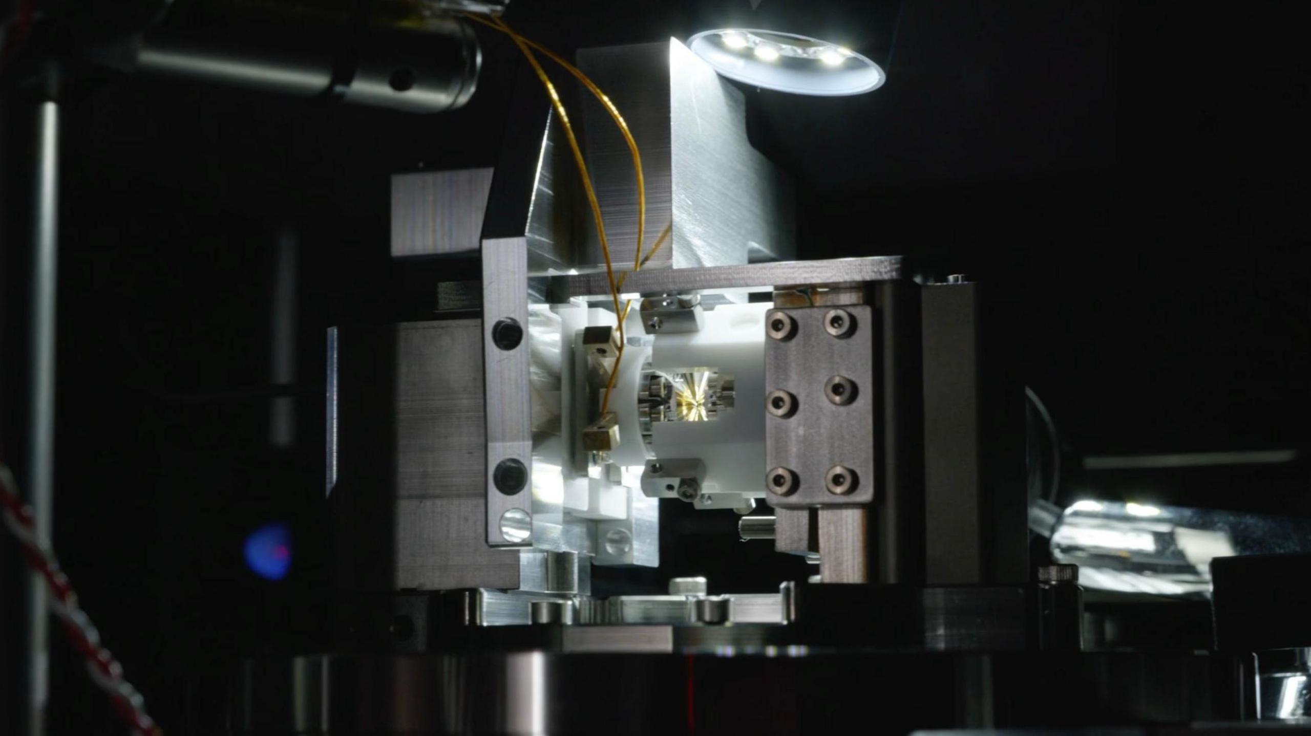 A qubit-photon interface. A piece of metallic hardware, lit by a single lamp in an otherwise dark room. In the centre of the image is a small metal and gold component, mounted inside white plastic, from which two wires emerge. The plastic is encased in a brushed metal surround. A lamp drops into the top of the image, shining light from the centre of the top of frame.