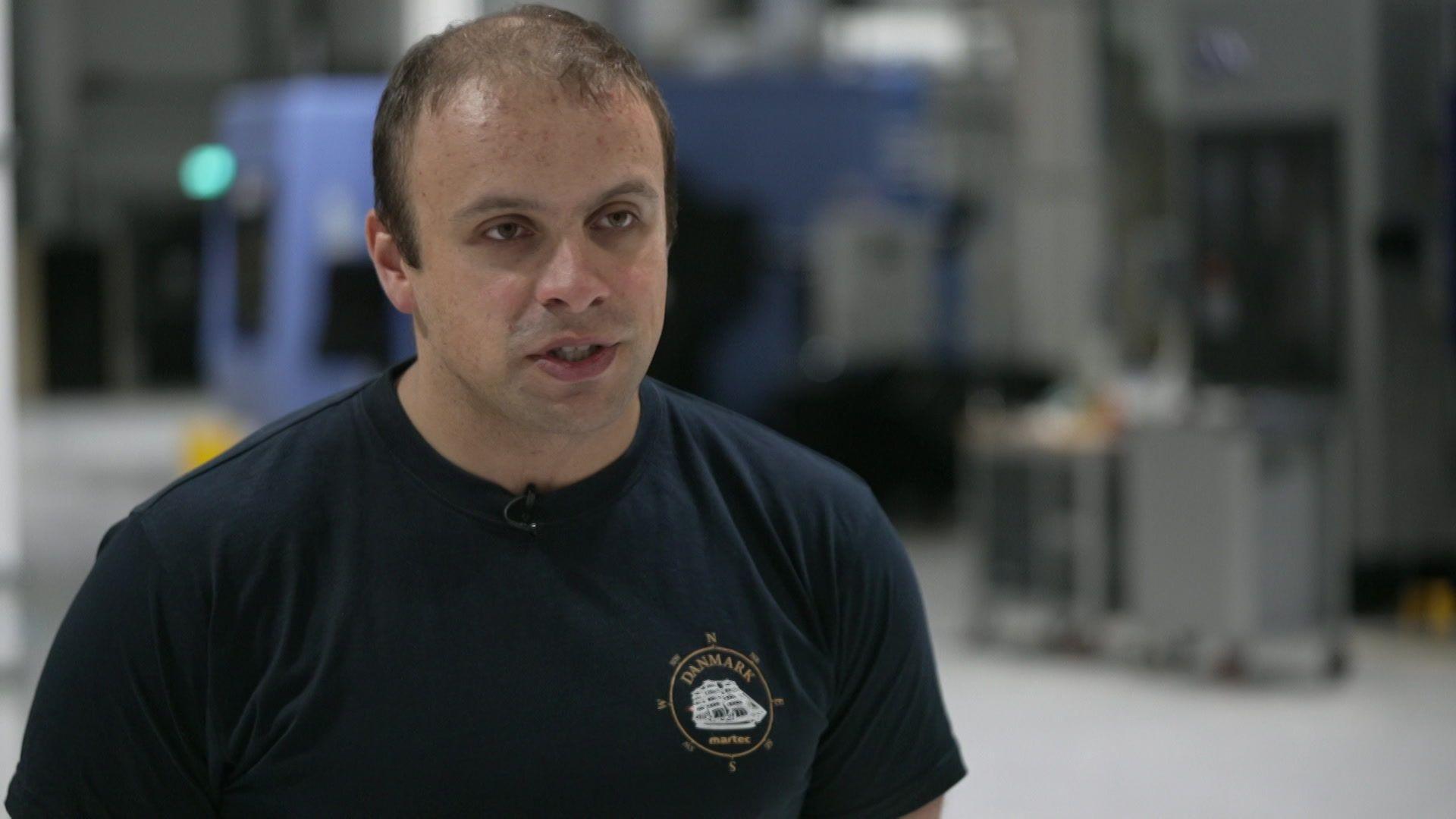 Alexander Fitzgerald, who is wearing a navy blue T-shirt and is standing in a large modern manufacturing space