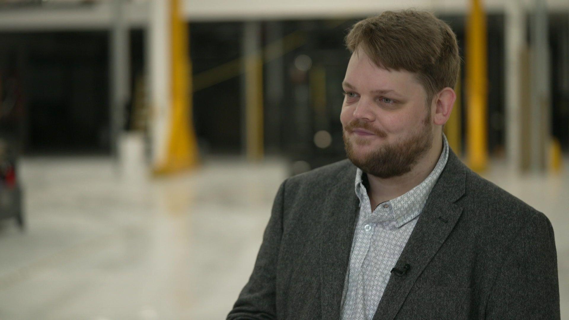 A man with fair hair and a beard wearing a tweed jacket and shirt standing in a factory