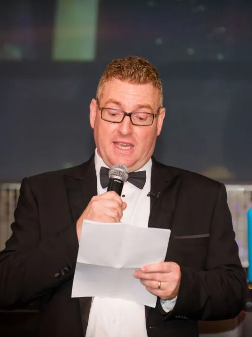 Man wearing a black-tie dinner suit stood by a microphone announcing winners at an awards event
