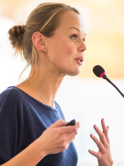 Photograph of a business woman on stage talking to a group of people at a conference