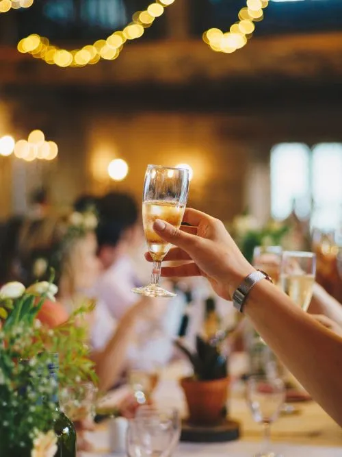 People sat at a table enjoying an event with a person holding up a champagne glass to the video camera