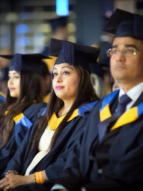 Several graduates sitting in the audience being filmed as they watch a graduation ceremony