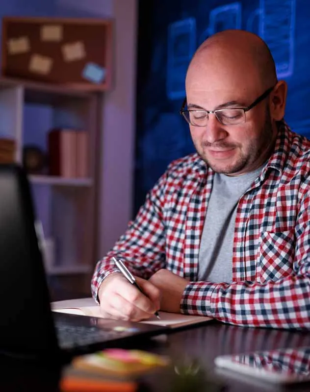 Mature man with checked shirt sitting a desk writing a script in a notebook