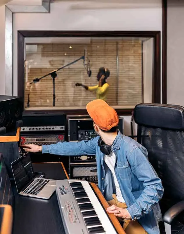 A woman singing in a recording booth being watched over by a sound engineer