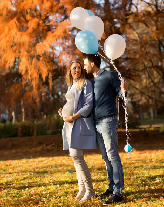 A pregnant woman and her partner standing in the park with blue and white balloons