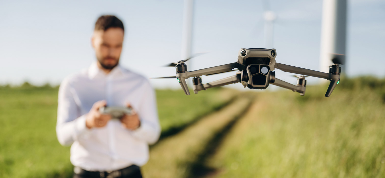 A man in a white shirt standing against holding the controller of a drone seen in foreground