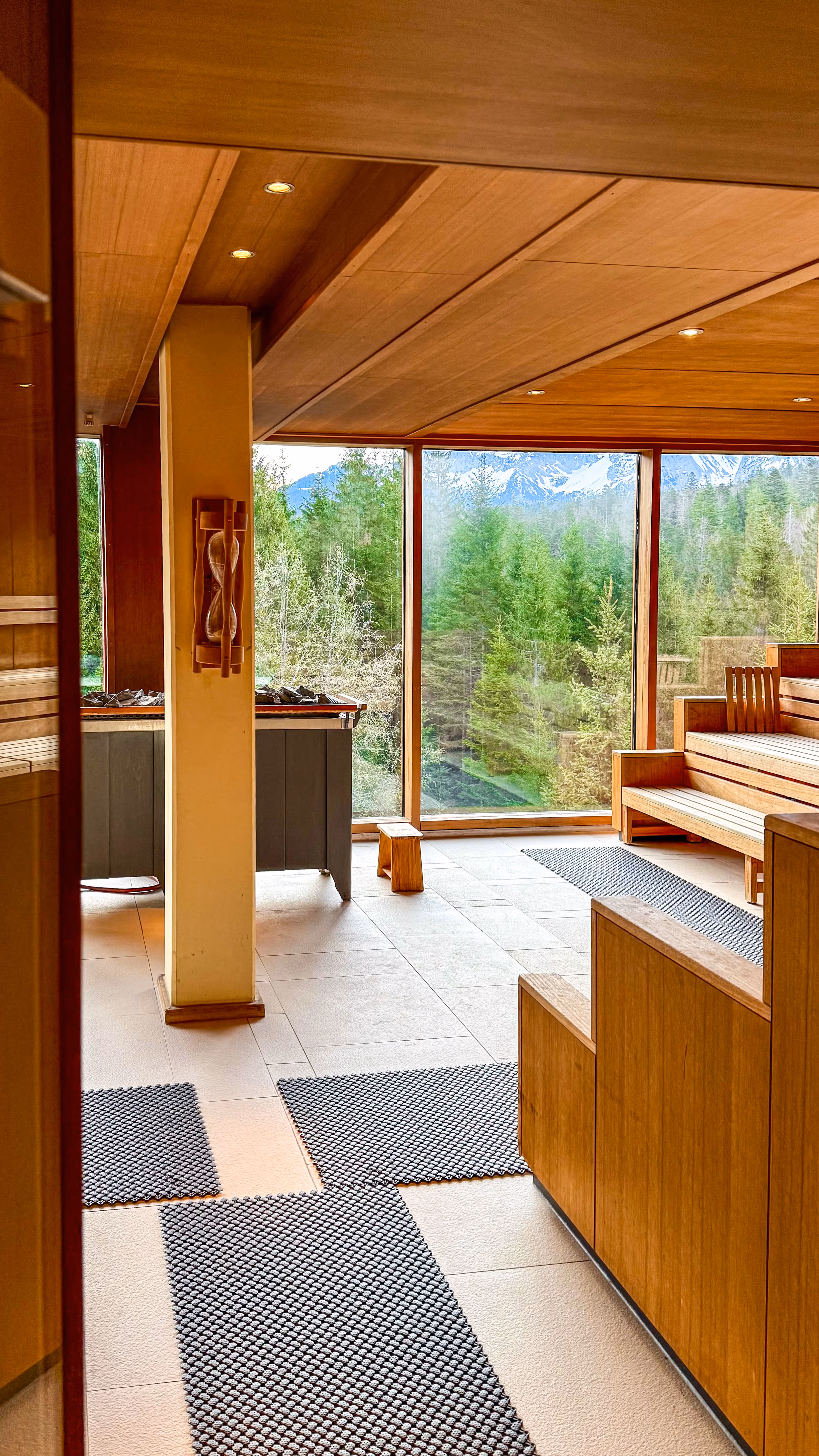 Indoor beige hammock chair with cushions near large glass windows overlooking an outdoor deck with lounge chairs and a mountain landscape.