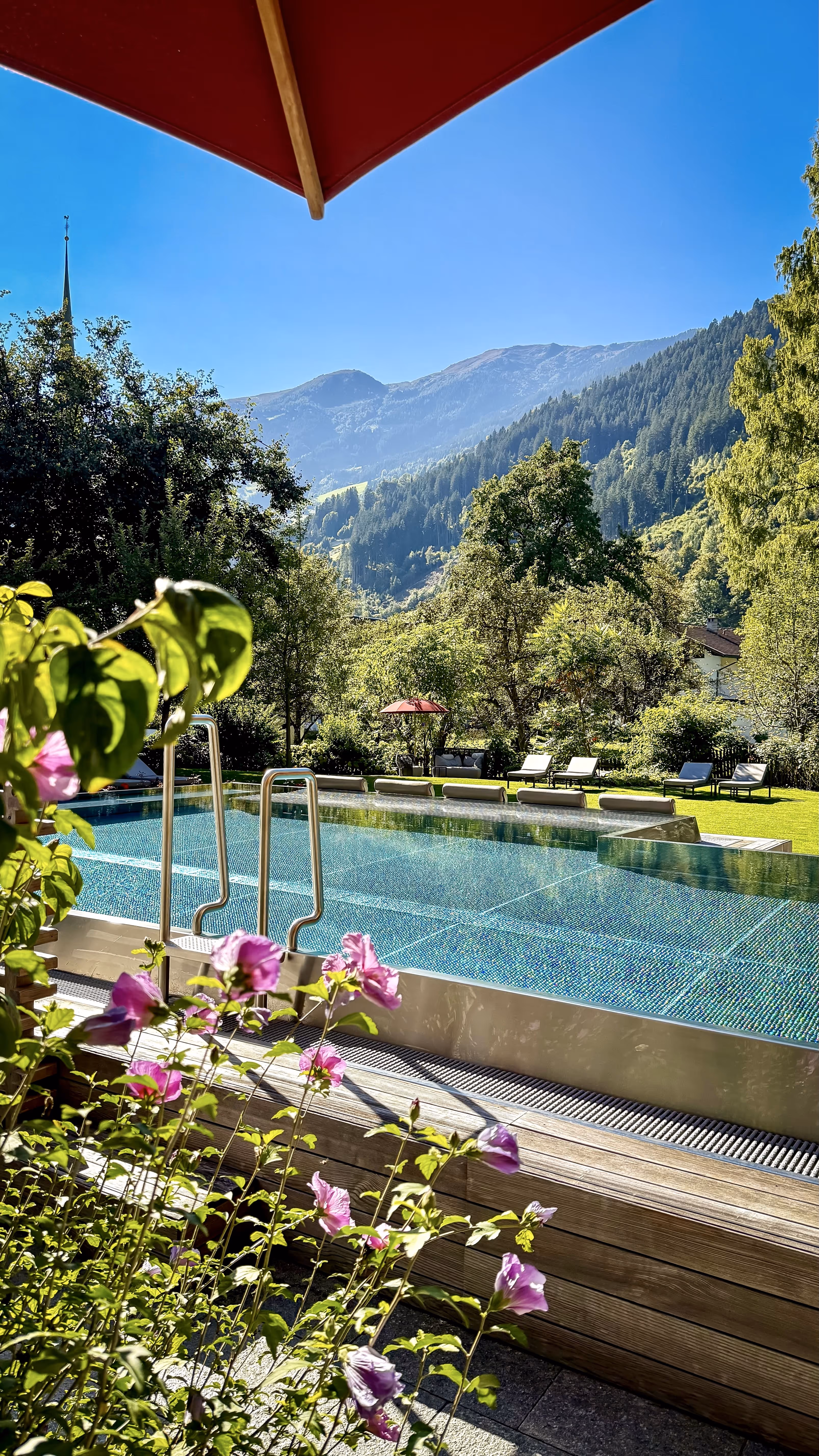 Outdoor swimming pool with clear water surrounded by green trees, pink flowers in foreground, lounge chairs on grass, and mountains under blue sky.
