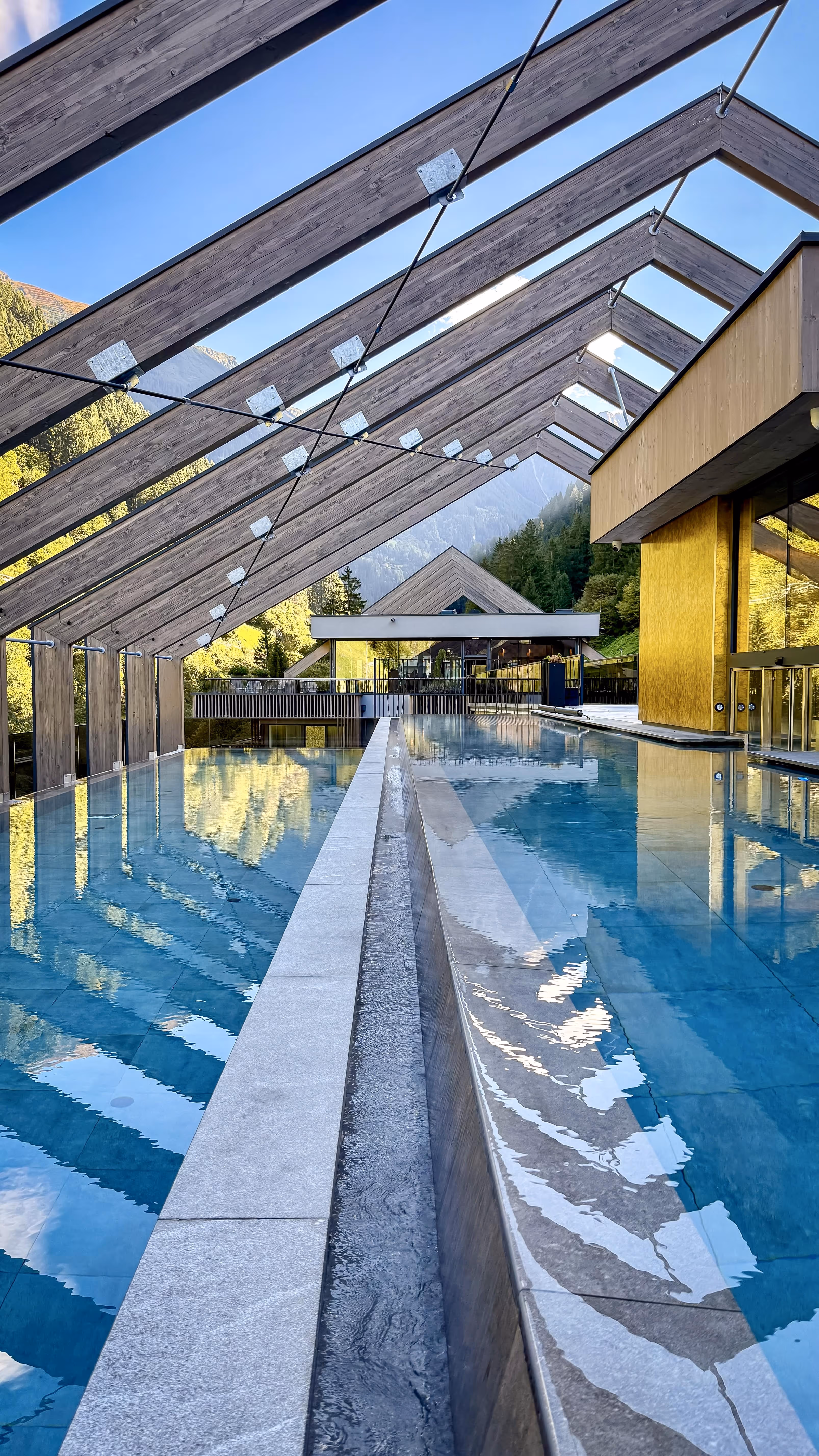 Modern outdoor pool with clear water under a wooden pergola structure set against a backdrop of green trees and mountains.