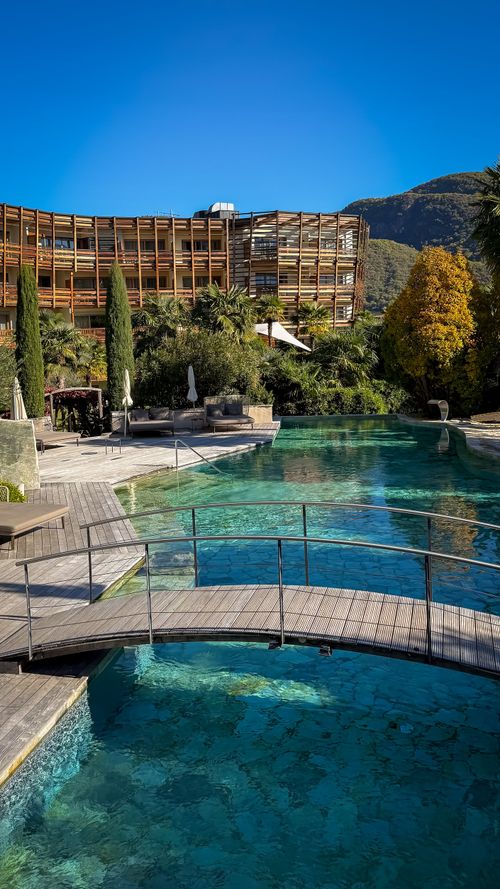 Outdoor pool with a small arched wooden bridge, surrounded by lounge chairs, greenery, and a hotel building with mountains in the background under clear blue sky.
