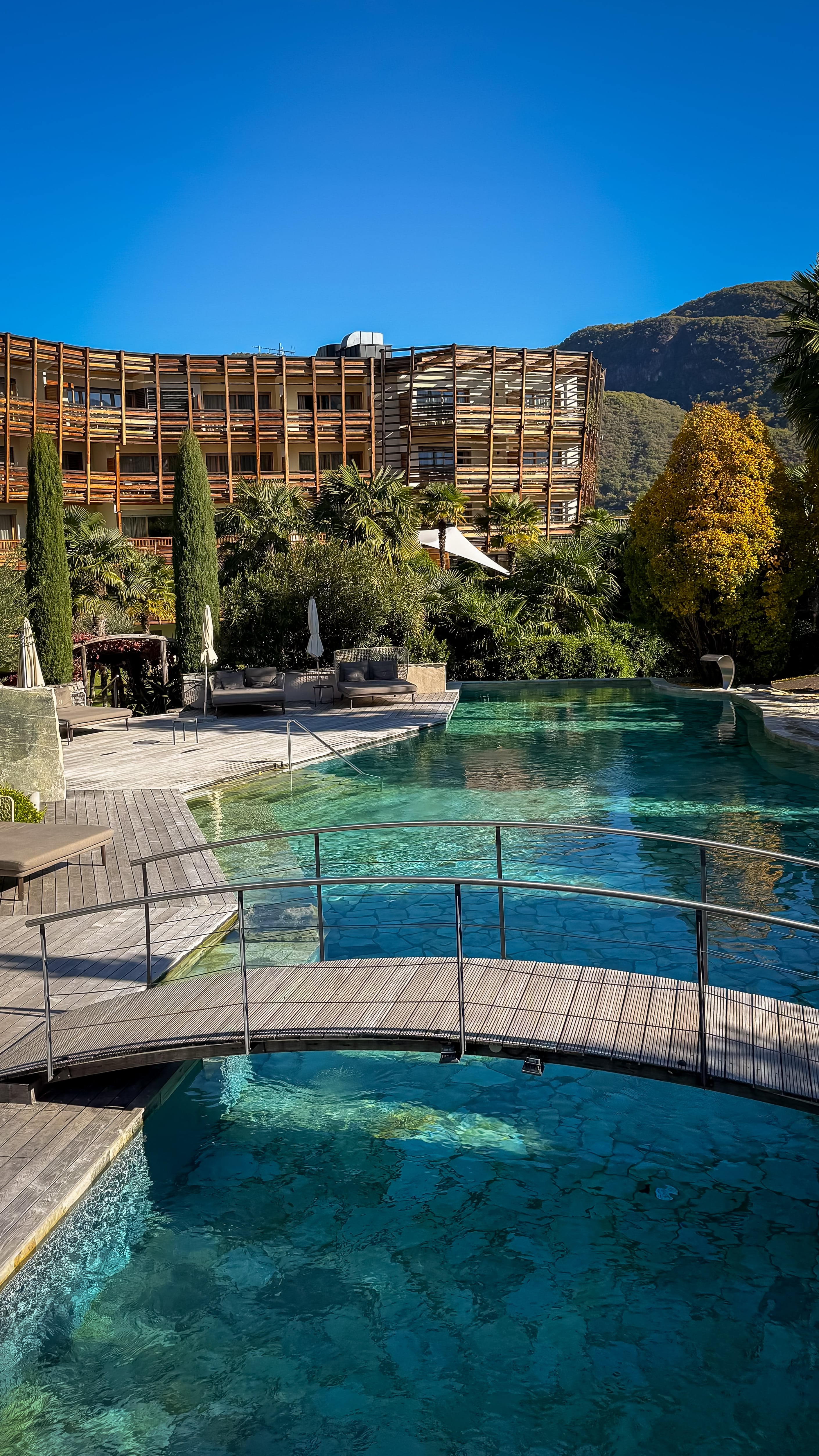 Outdoor pool with a small arched wooden bridge, surrounded by lounge chairs, greenery, and a hotel building with mountains in the background under clear blue sky.