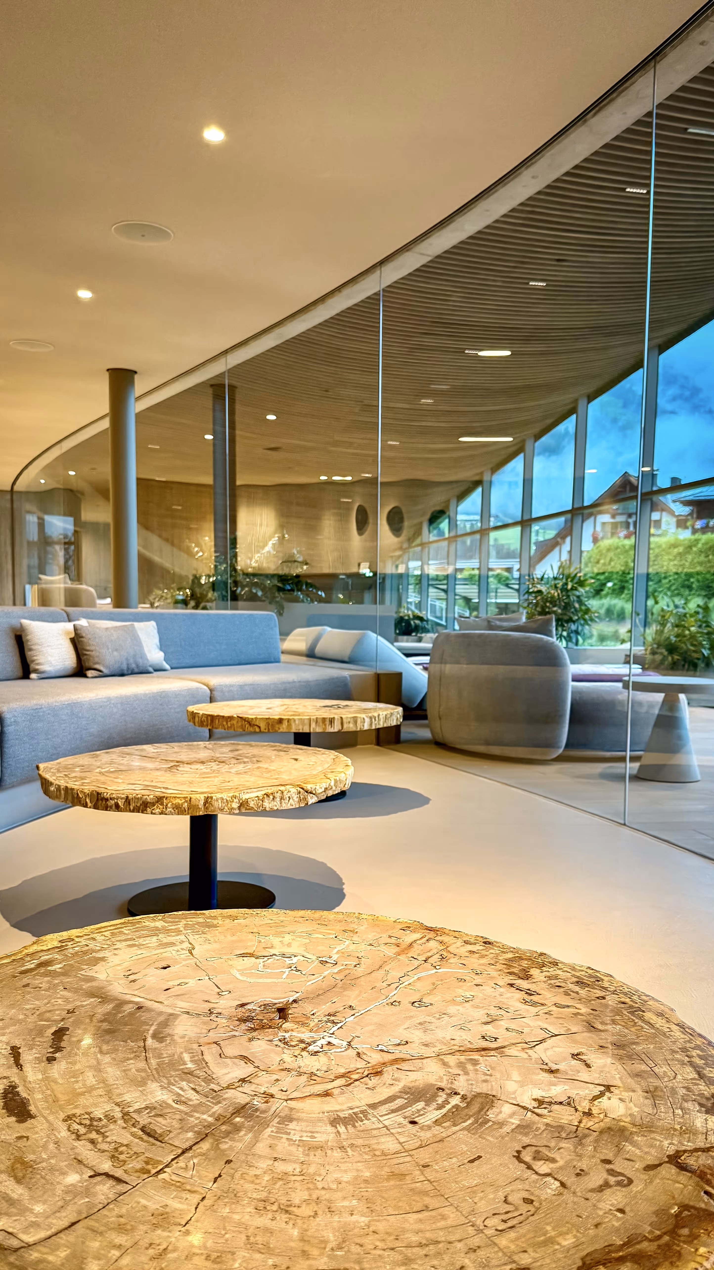 Modern lounge with round wooden tables, gray sofas, large glass windows, and views of greenery outside.
