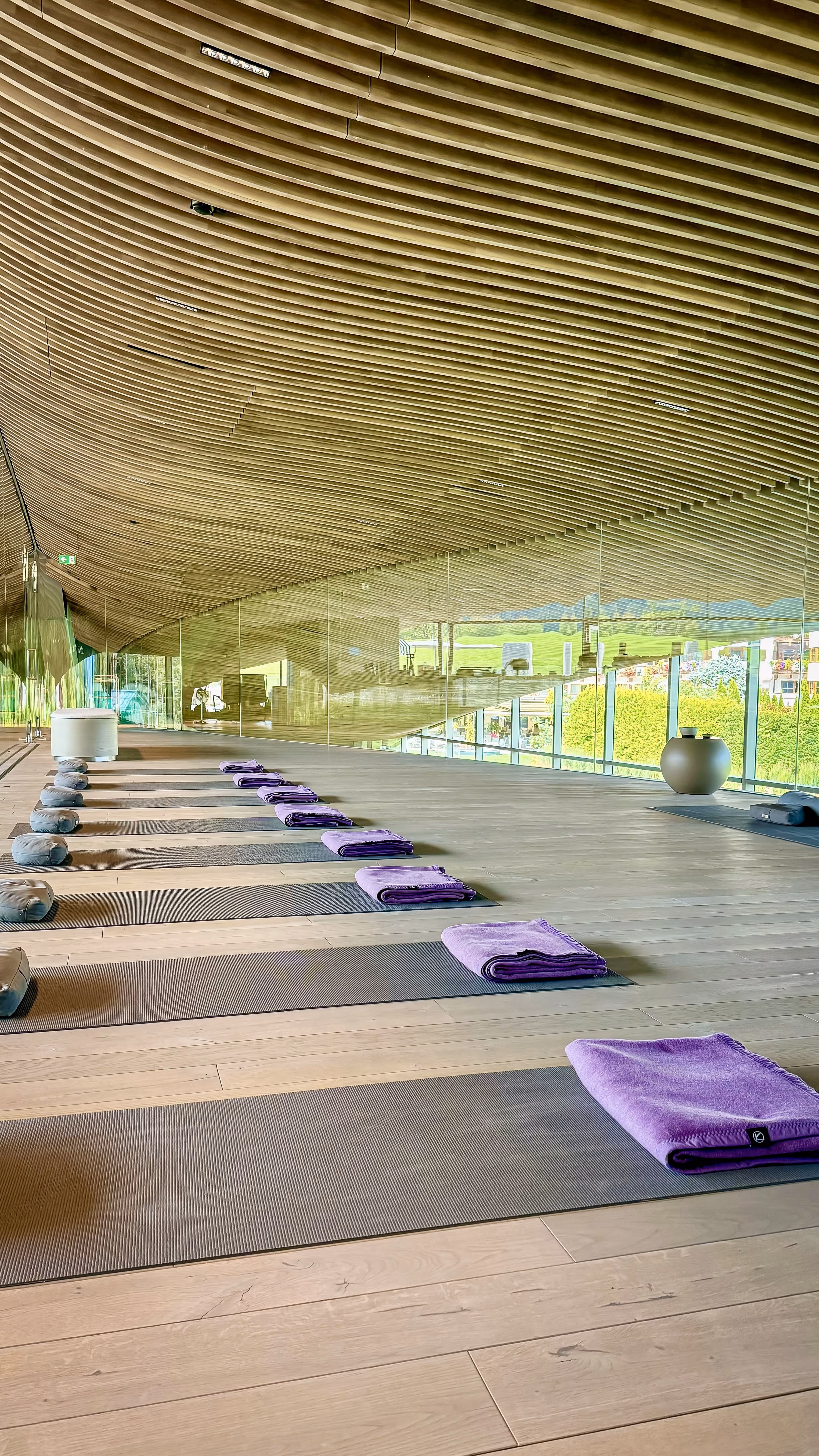 Modern yoga studio with wooden slatted ceiling, lined mats each with a purple towel and cushion, and floor-to-ceiling windows overlooking greenery.