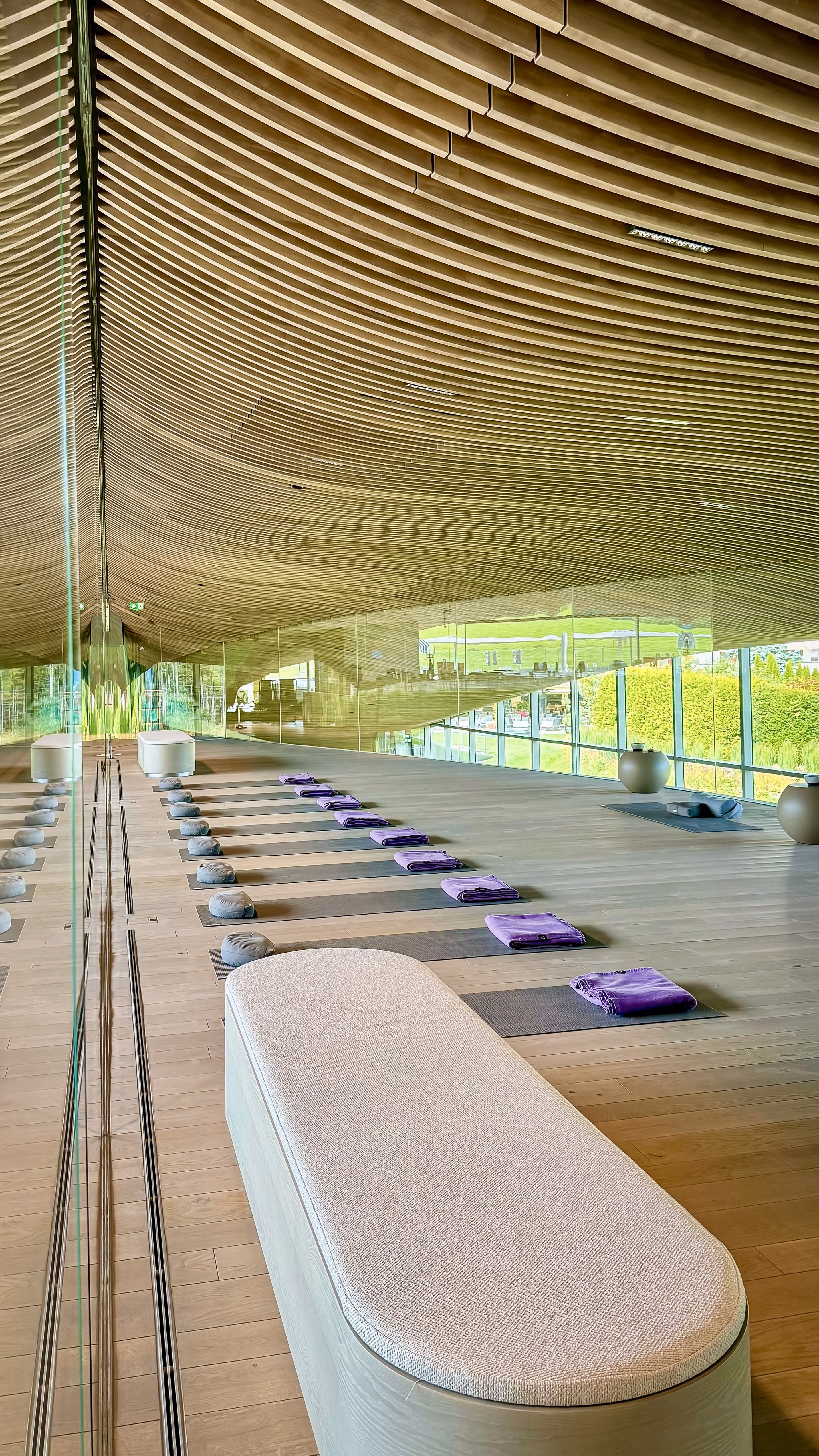 Modern indoor seating area with two plush gray armchairs, a round wooden table with a black base, and large green plants under a wooden slatted ceiling.