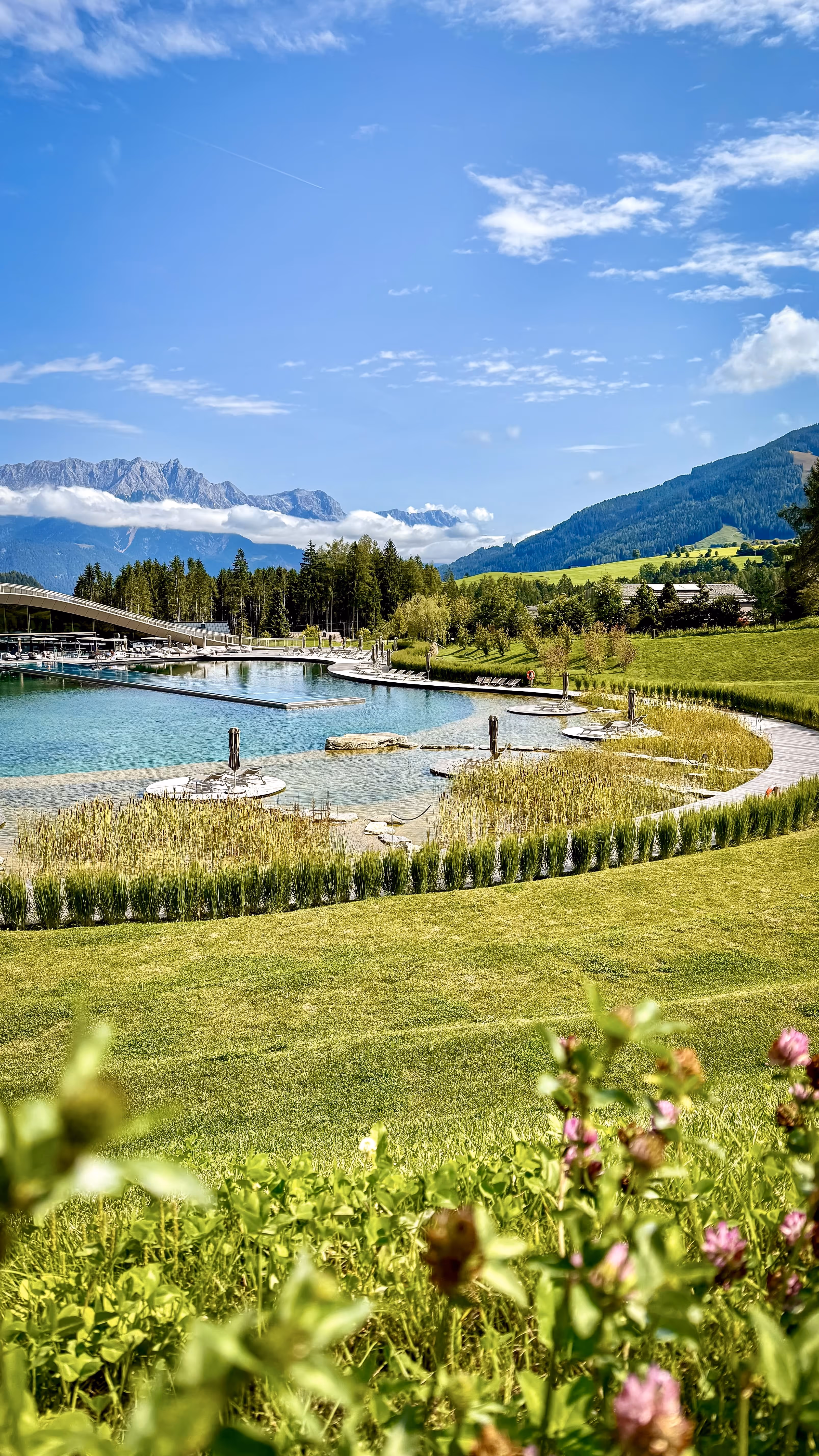 Natural swimming pool with clear water surrounded by greenery and mountains under a partly cloudy blue sky.