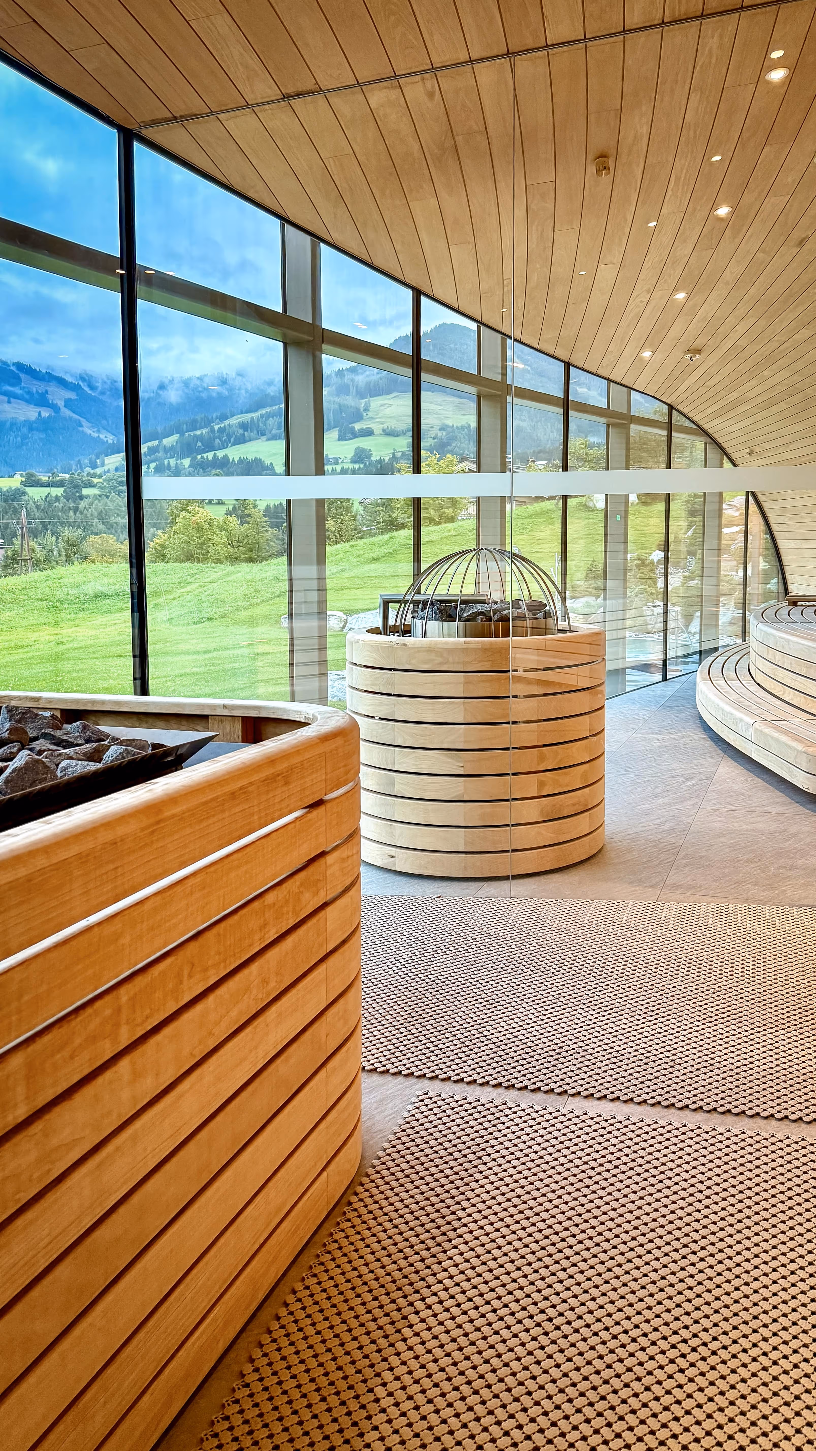 Modern indoor sauna with curved wooden benches, stone heaters, and large windows showing a green mountainous landscape.