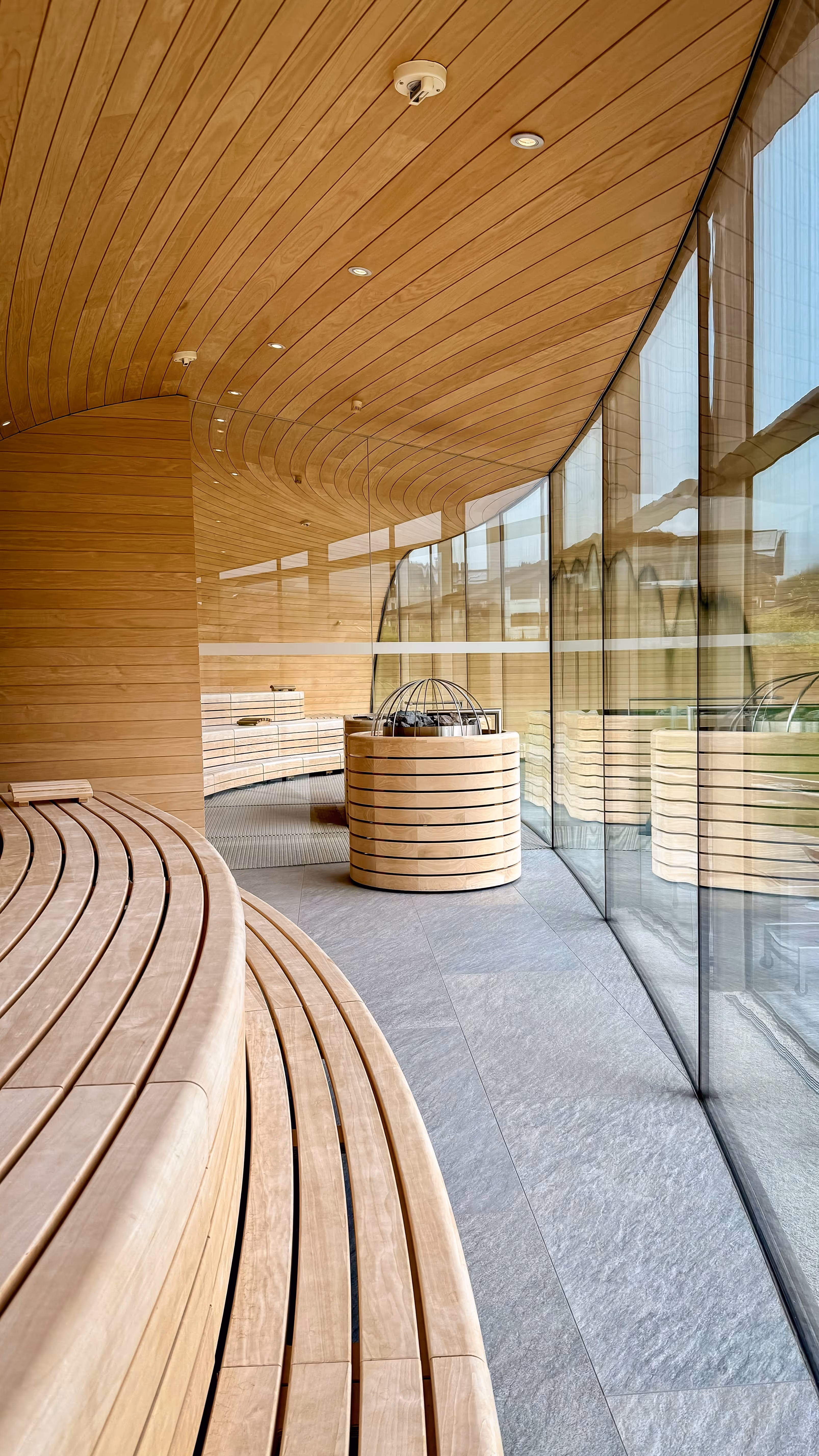 Curved wooden sauna interior with slatted wooden bench and a small wooden backrest, next to a large arched window showing green grass and an outdoor pool.