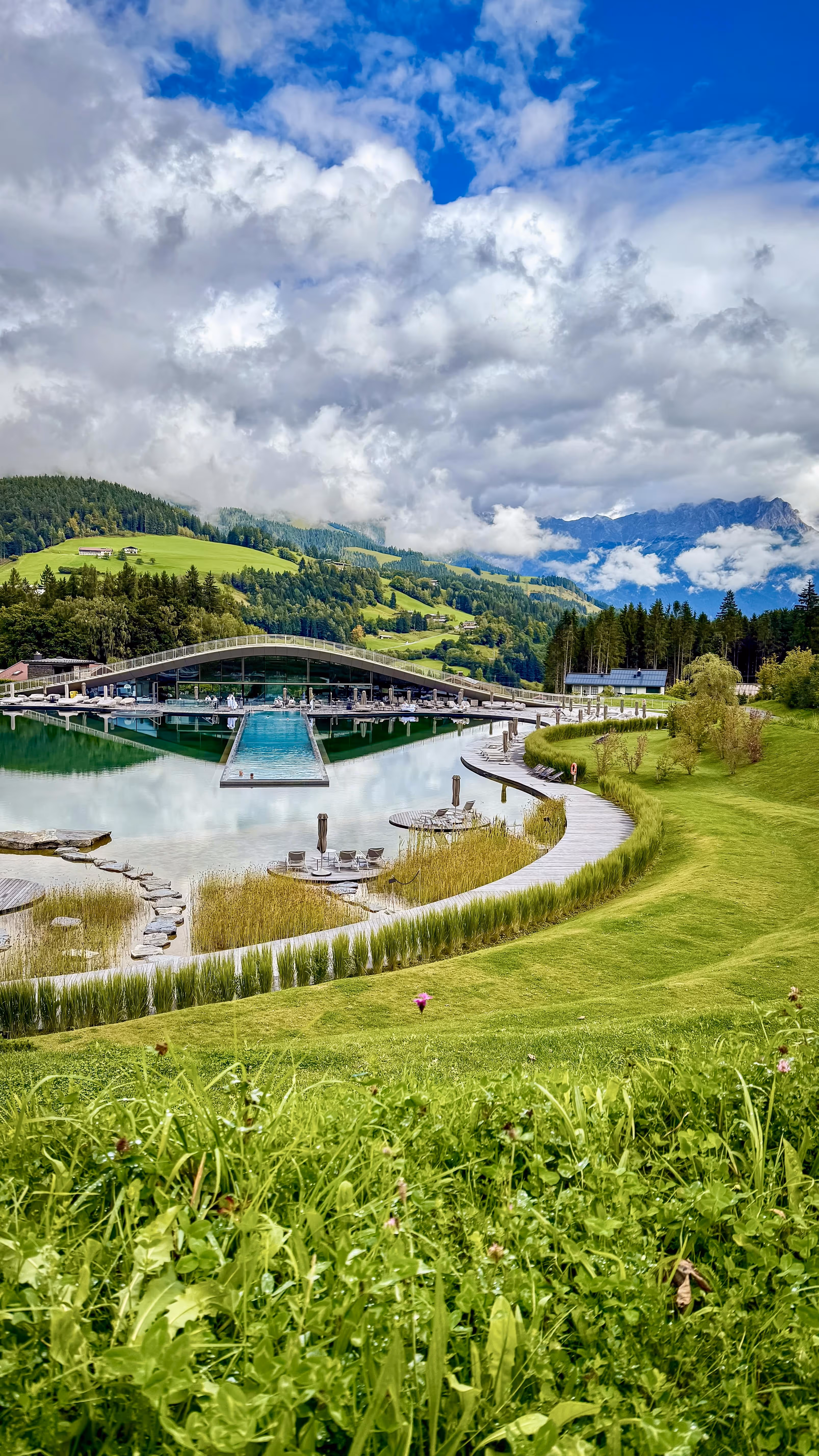 Modern building with a wave-shaped roof beside a reflective lake surrounded by green hills and forest under a partly cloudy blue sky.