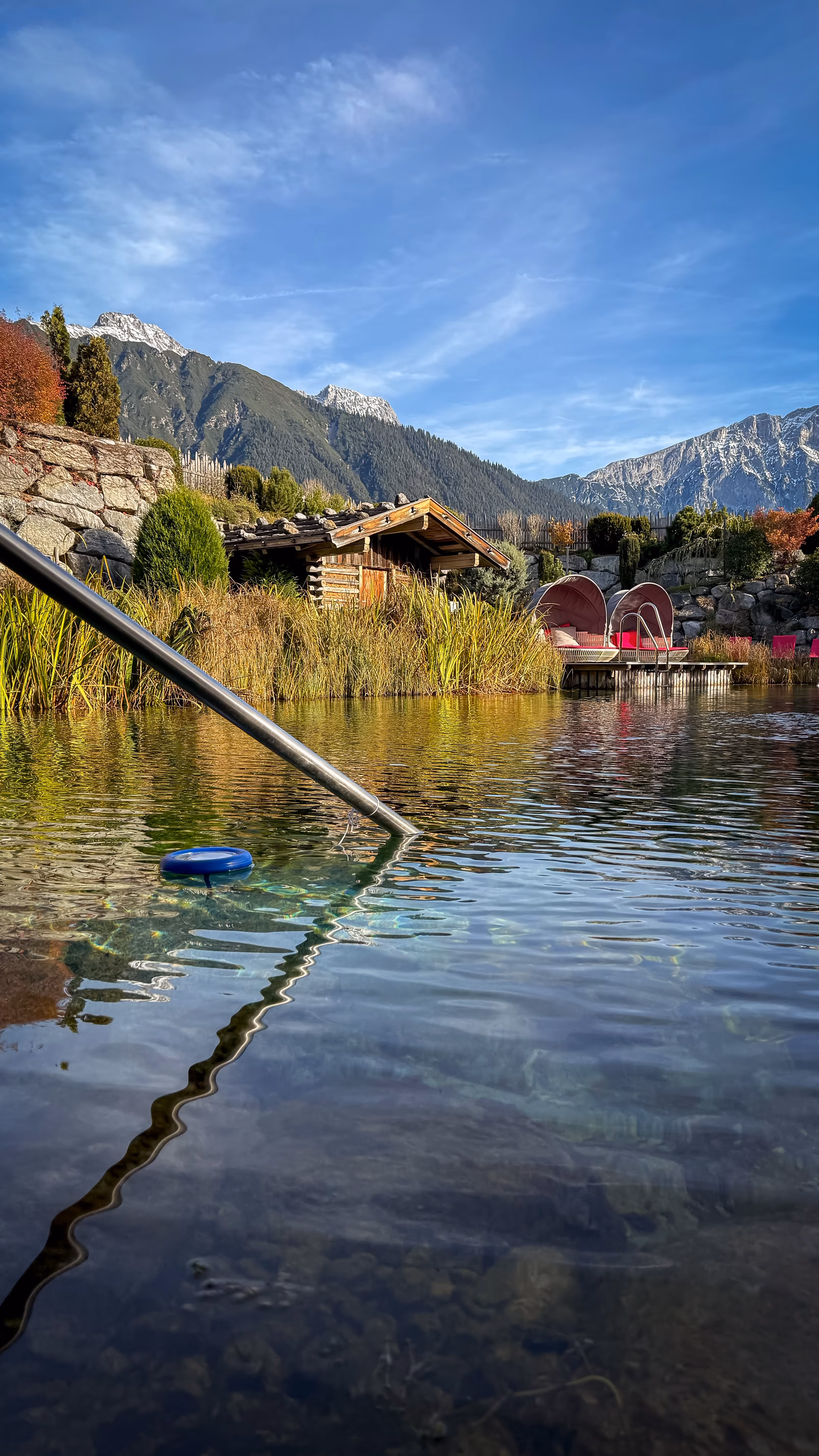 Clear natural pond with plants and a wooden cabin in the background, snow-capped mountains under a blue sky.