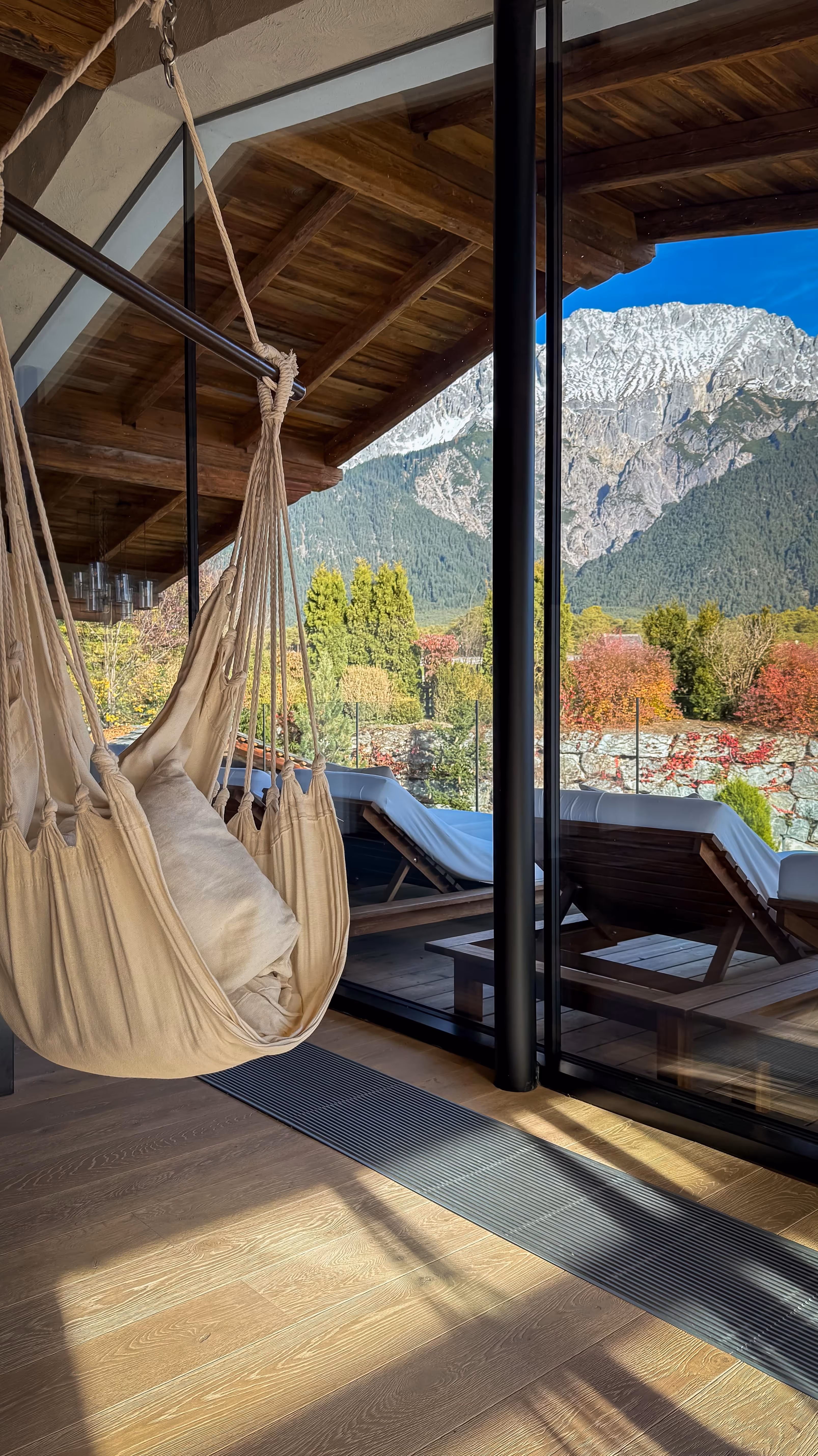 Indoor beige hammock chair with cushions near large glass windows overlooking an outdoor deck with lounge chairs and a mountain landscape.