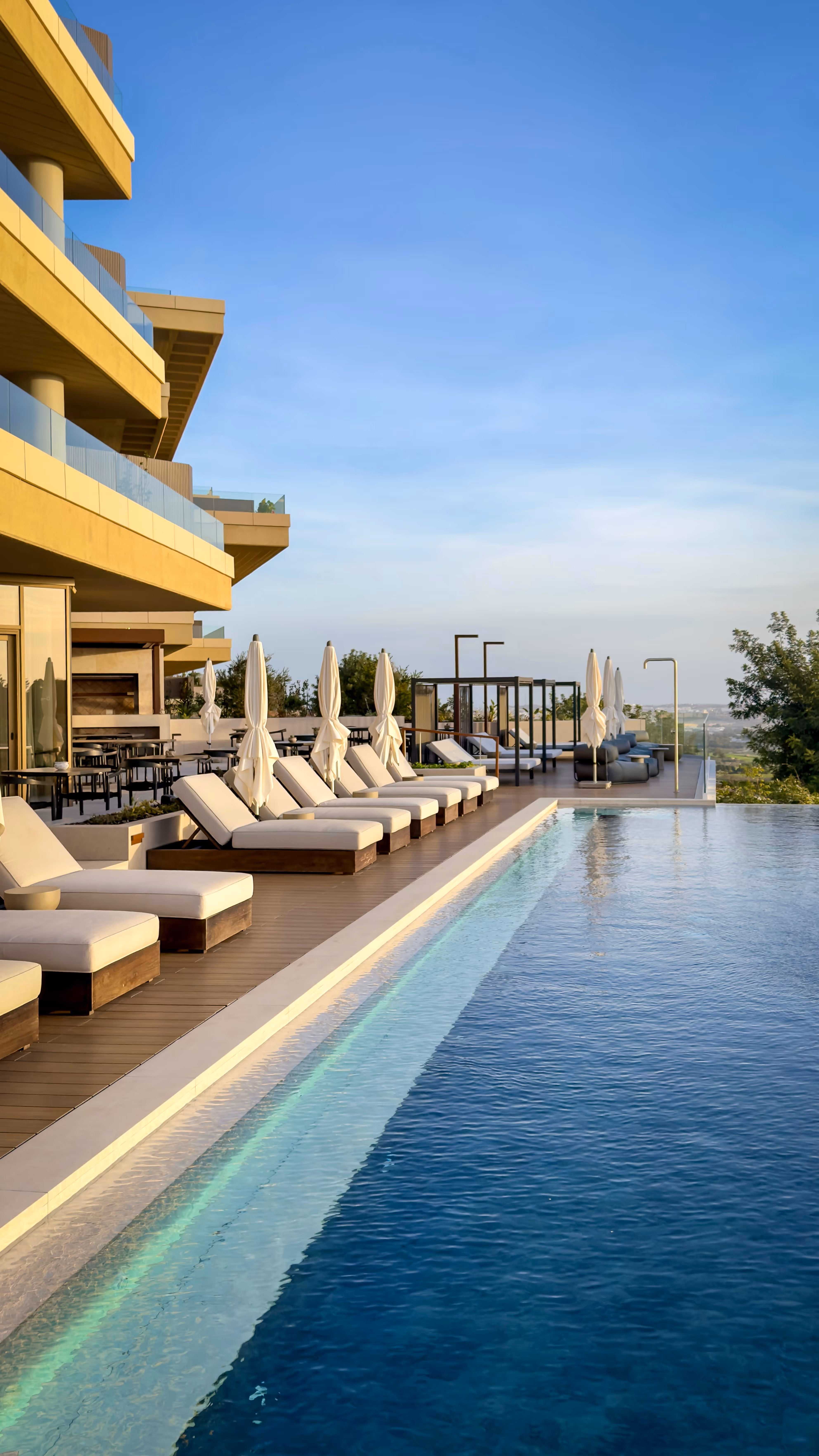Modern lounge area with natural wood round coffee tables, light gray sofas, and large glass windows revealing greenery outside.