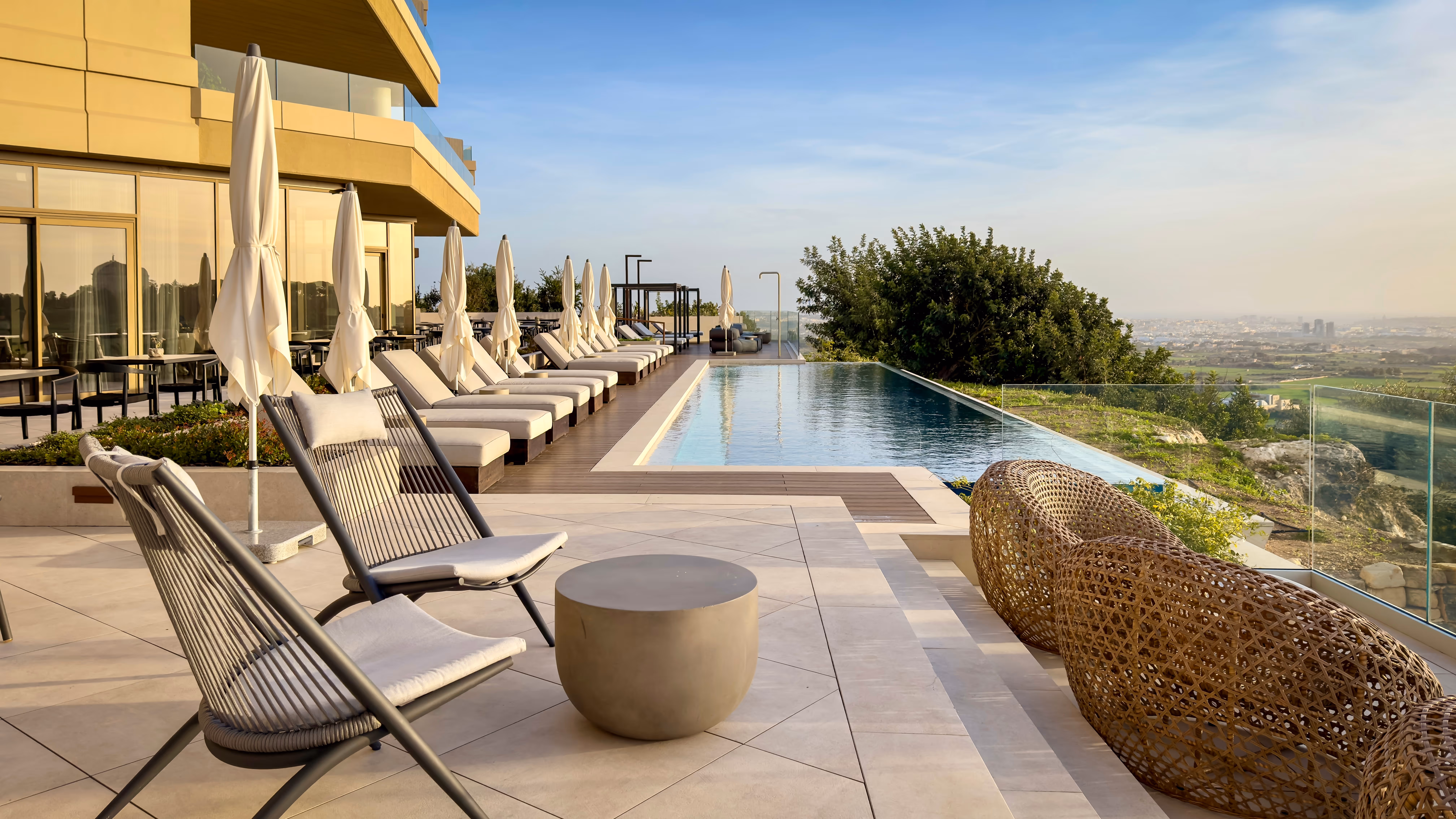 Outdoor pool beside green lawn with red flowering bushes, lounge chairs, and mountainous forest background under clear blue sky.