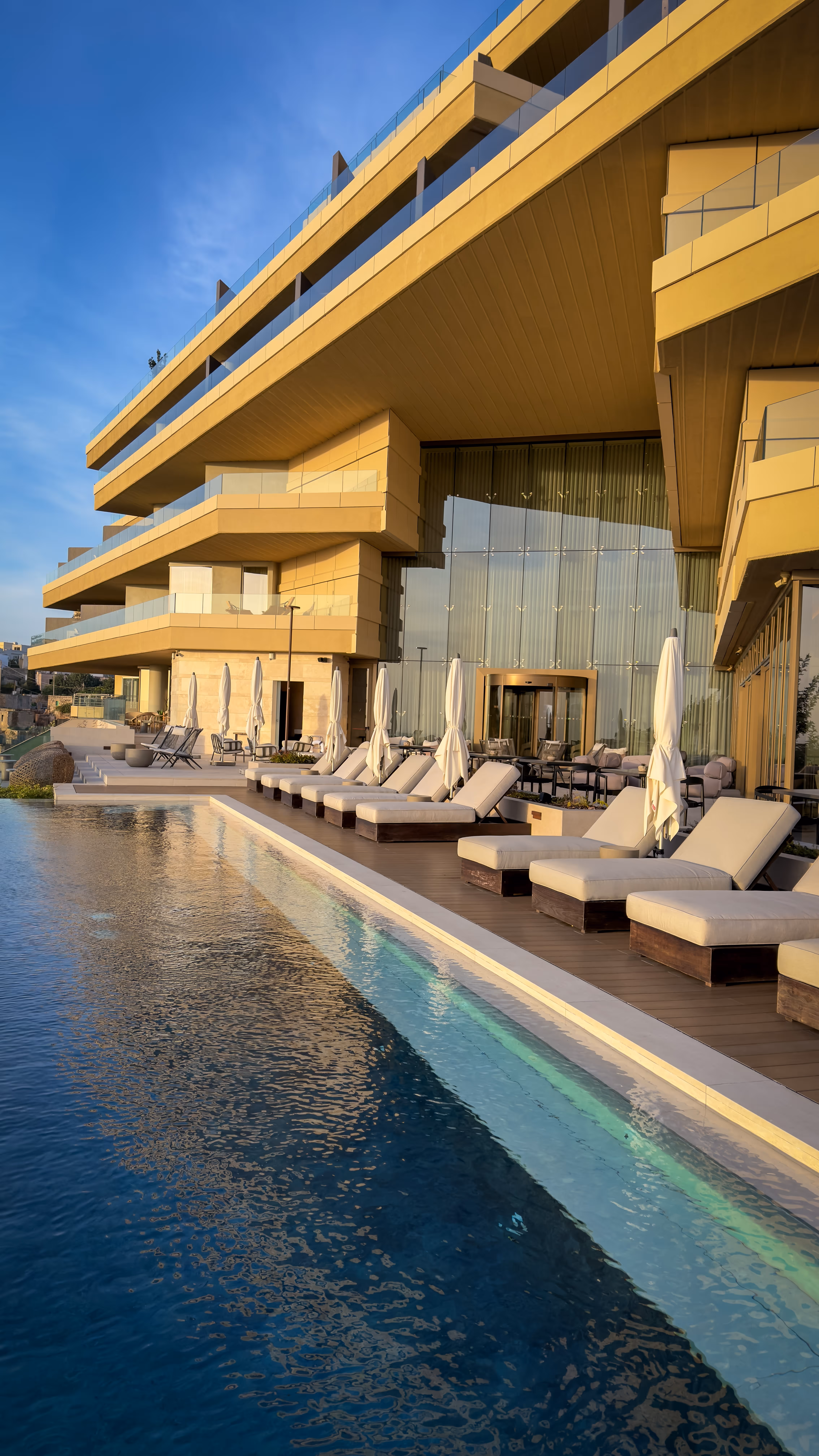 Clear natural swimming pond with lounge chairs and wooden decks, mountains and blue sky in the background.