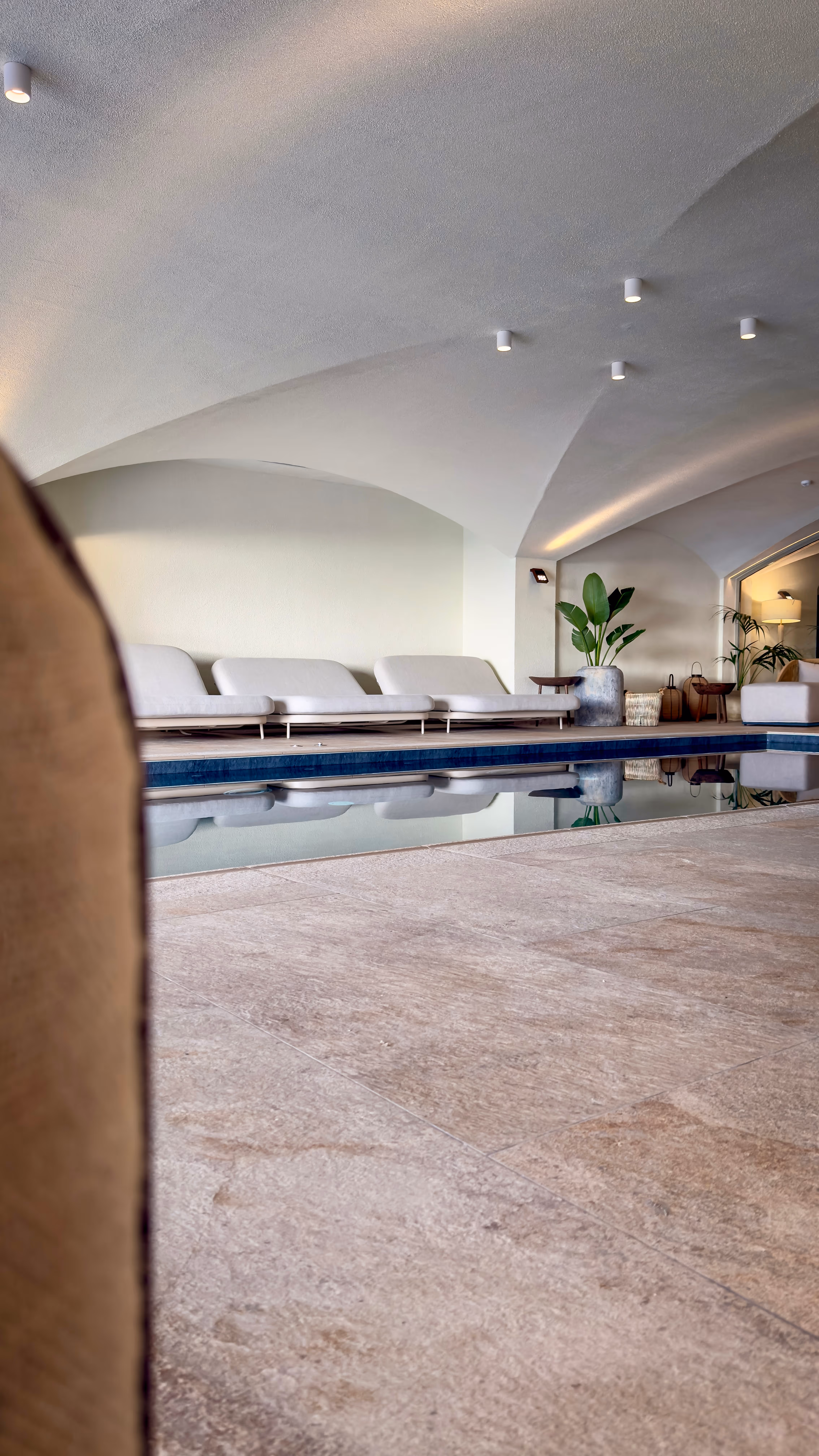 Indoor pool area with beige lounge chairs, arched ceiling, and potted plants near the water.