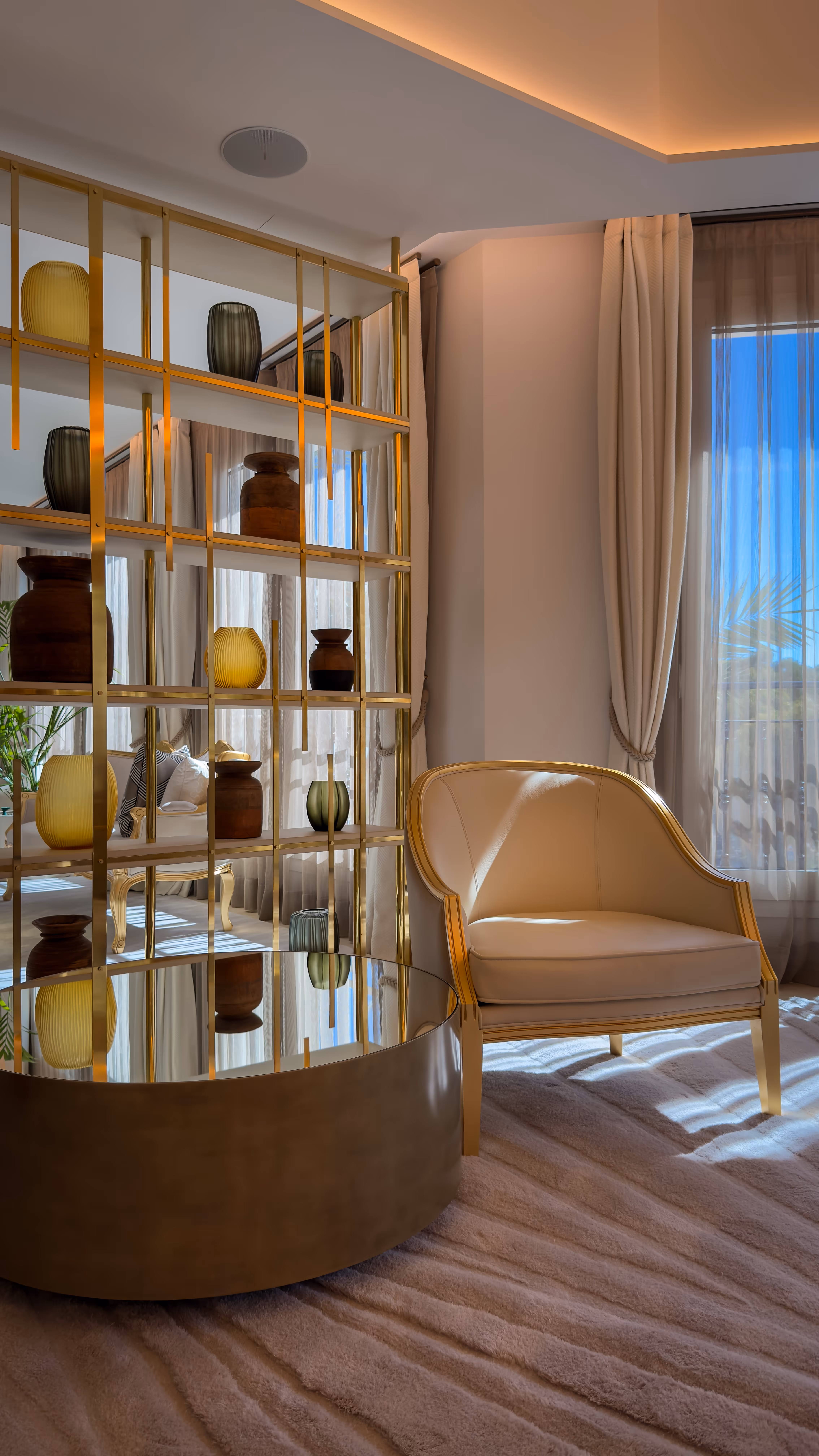 Elegant beige armchair next to a gold and mirrored shelving unit with decorative vases in a sunlit room with floor-to-ceiling curtains.