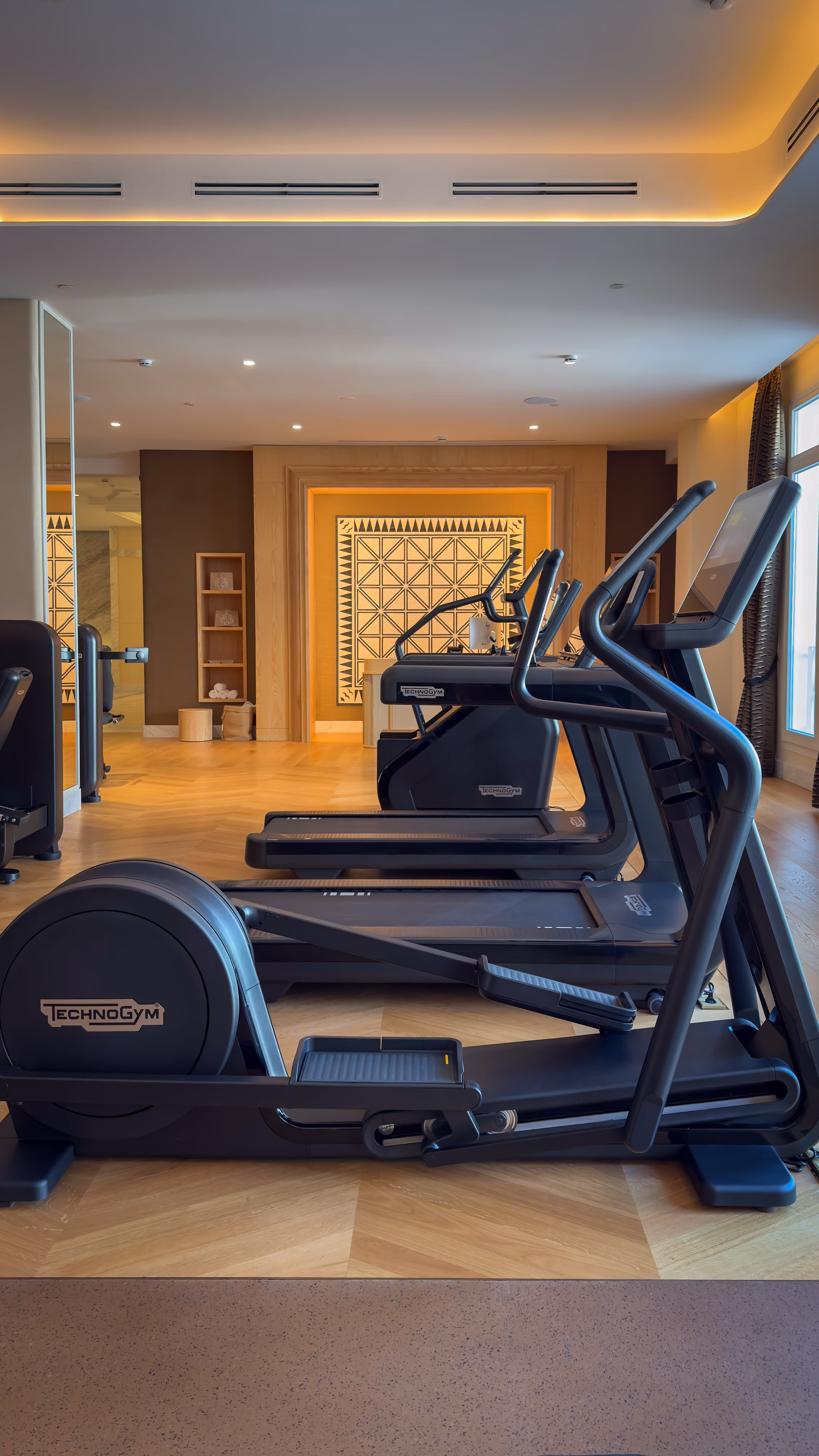 Modern gym room with black Technogym treadmills lined up on a wooden floor facing a decorative wall panel.