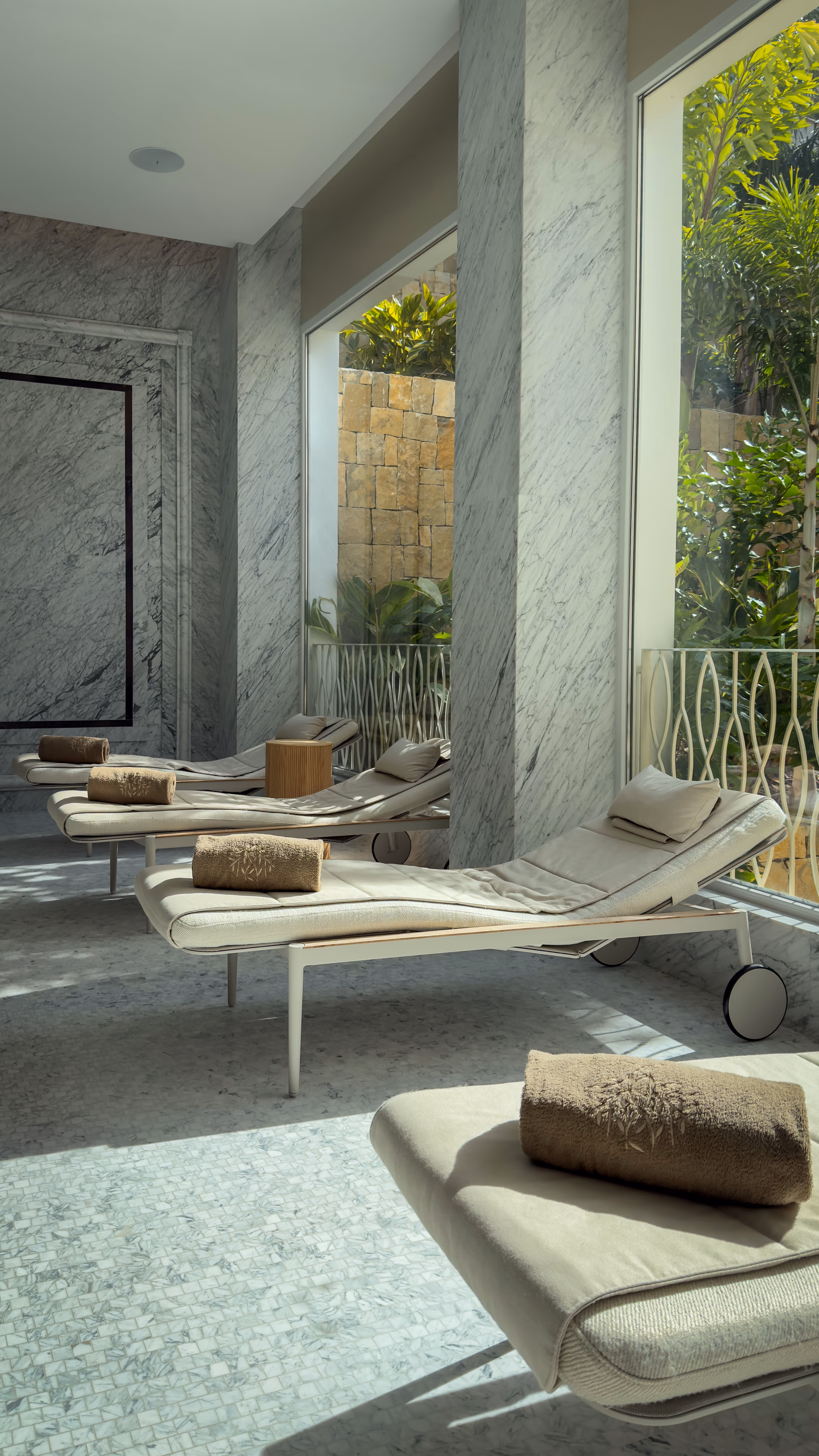 Indoor beige hammock chair with cushions near large glass windows overlooking an outdoor deck with lounge chairs and a mountain landscape.