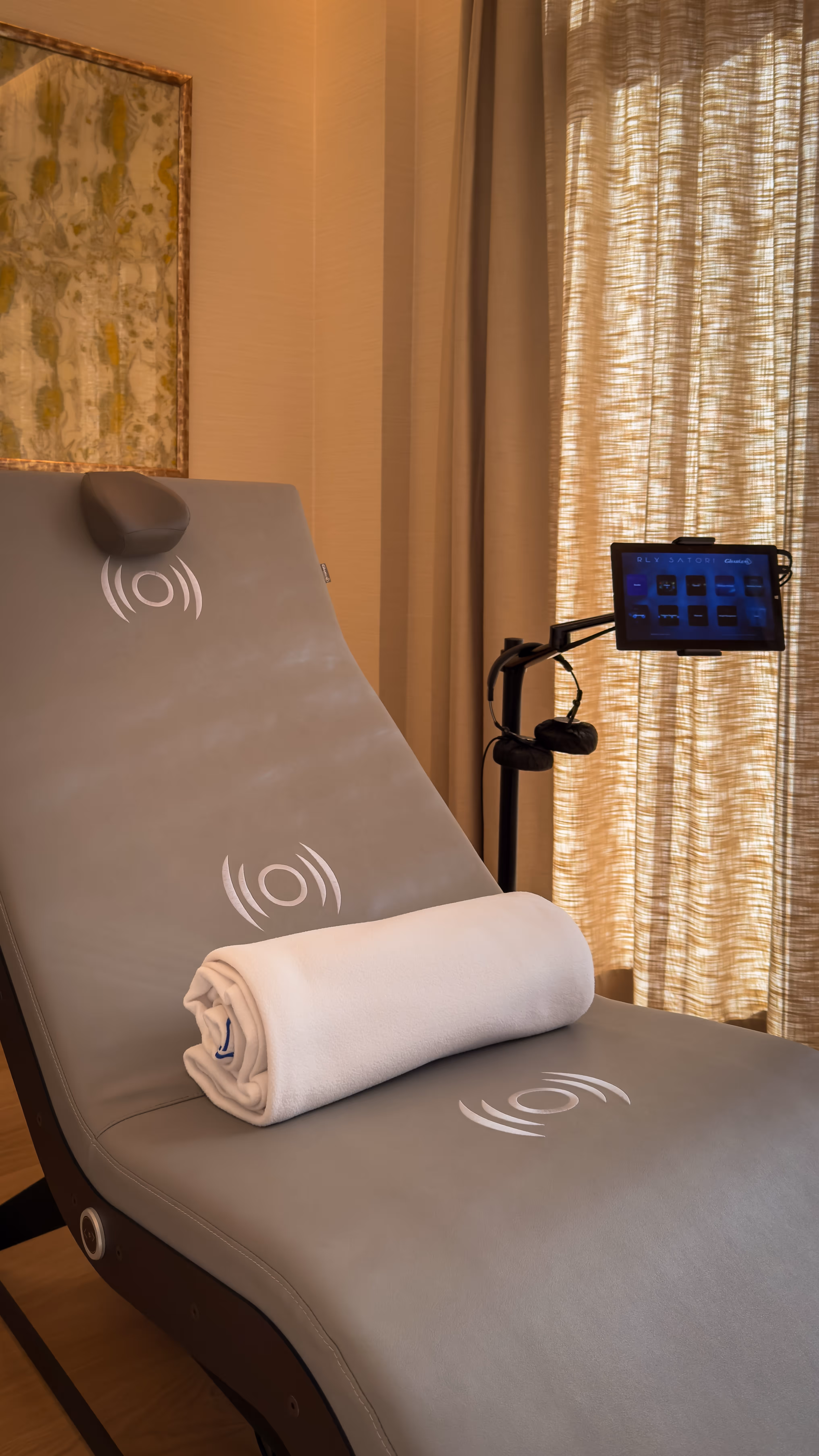 Modern massage chair with a rolled white towel, headset, and control screen in a softly lit room with beige curtains.