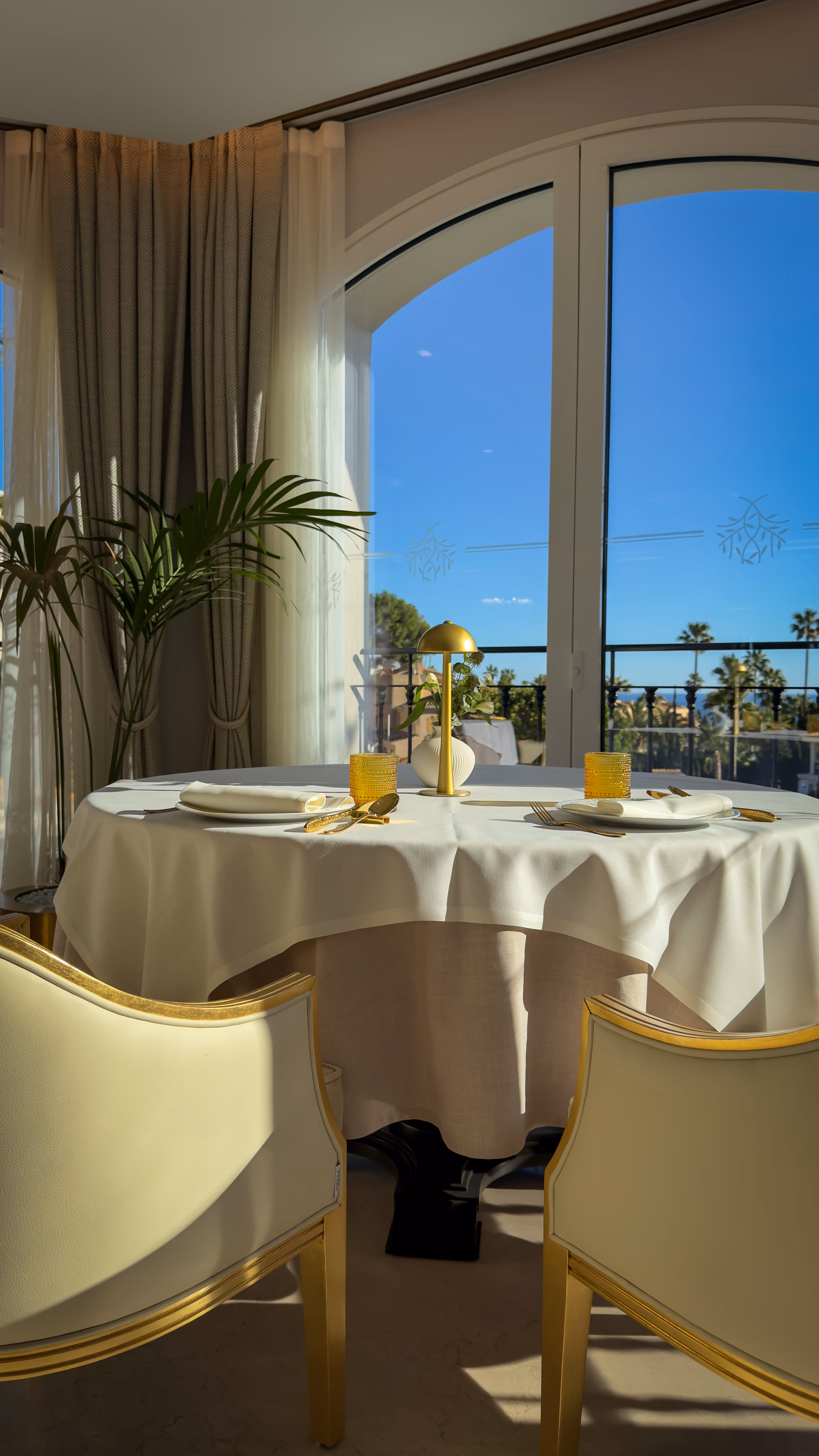 Elegant dining table set for two with golden cutlery, amber glasses, a small lamp, and a flower vase near large arched windows with a sunny outdoor view.