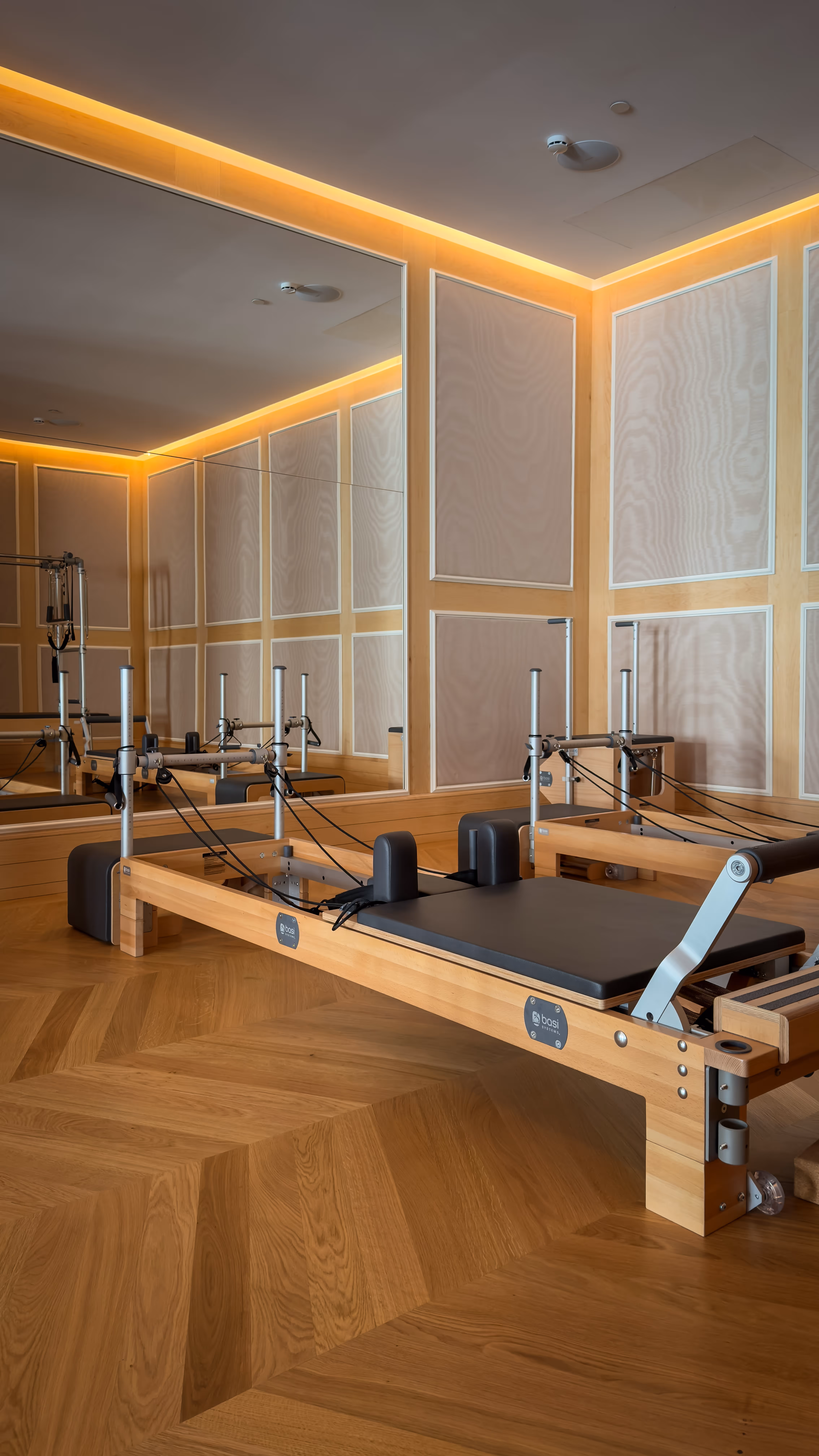 Wooden Pilates reformer machine with black cushioning in a well-lit studio with mirrored walls and wood paneling.