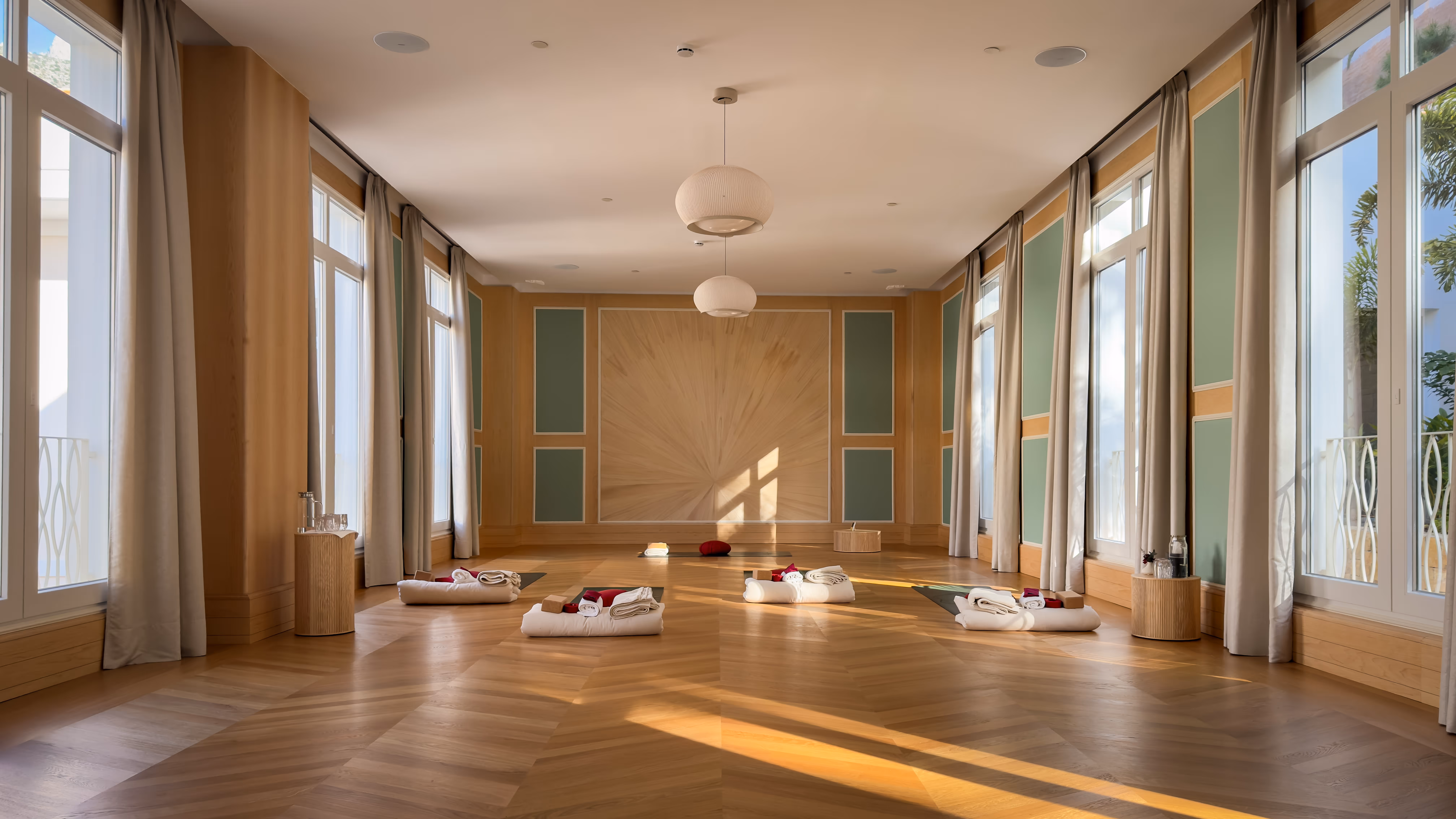 Bright yoga studio with wooden floors, large windows with curtains, ceiling lights, and yoga mats with cushions and towels arranged on the floor.