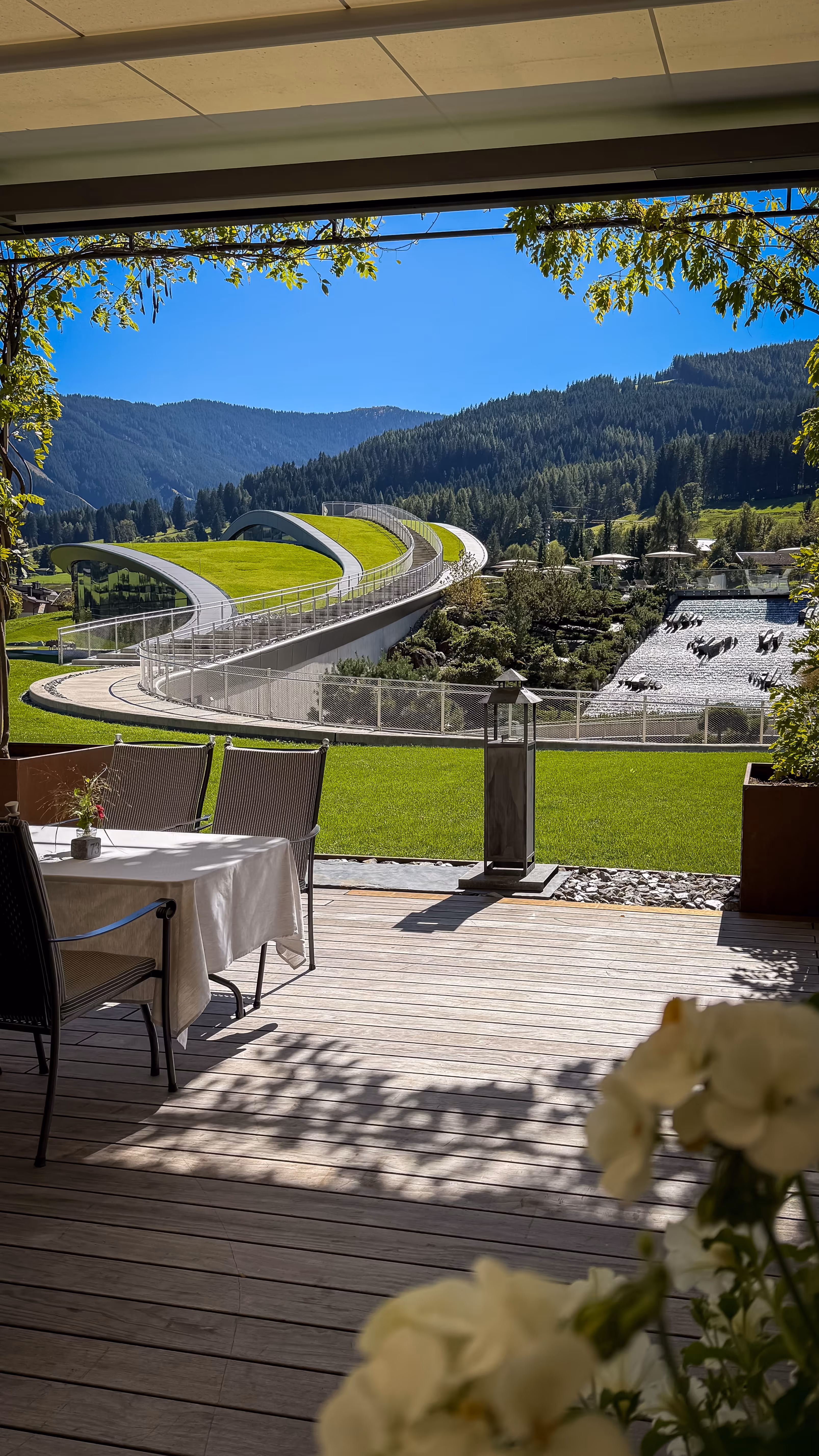 Patio with a table and chairs overlooking a modern building with green curved roofs, surrounded by grass, trees, and mountains under a clear blue sky.
