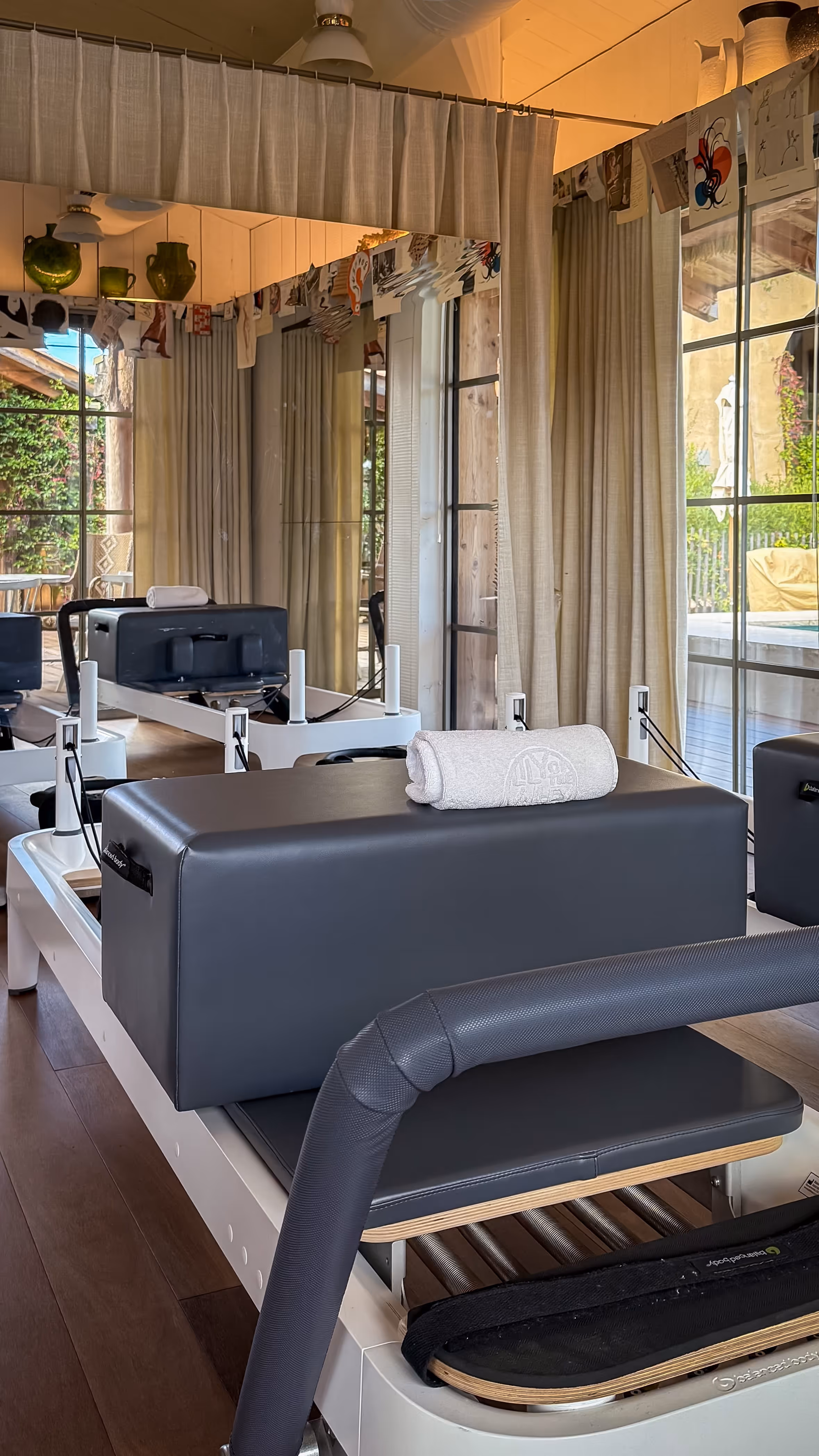 Pilates reformer machine with a rolled towel on top in a bright exercise room with large windows and curtains.