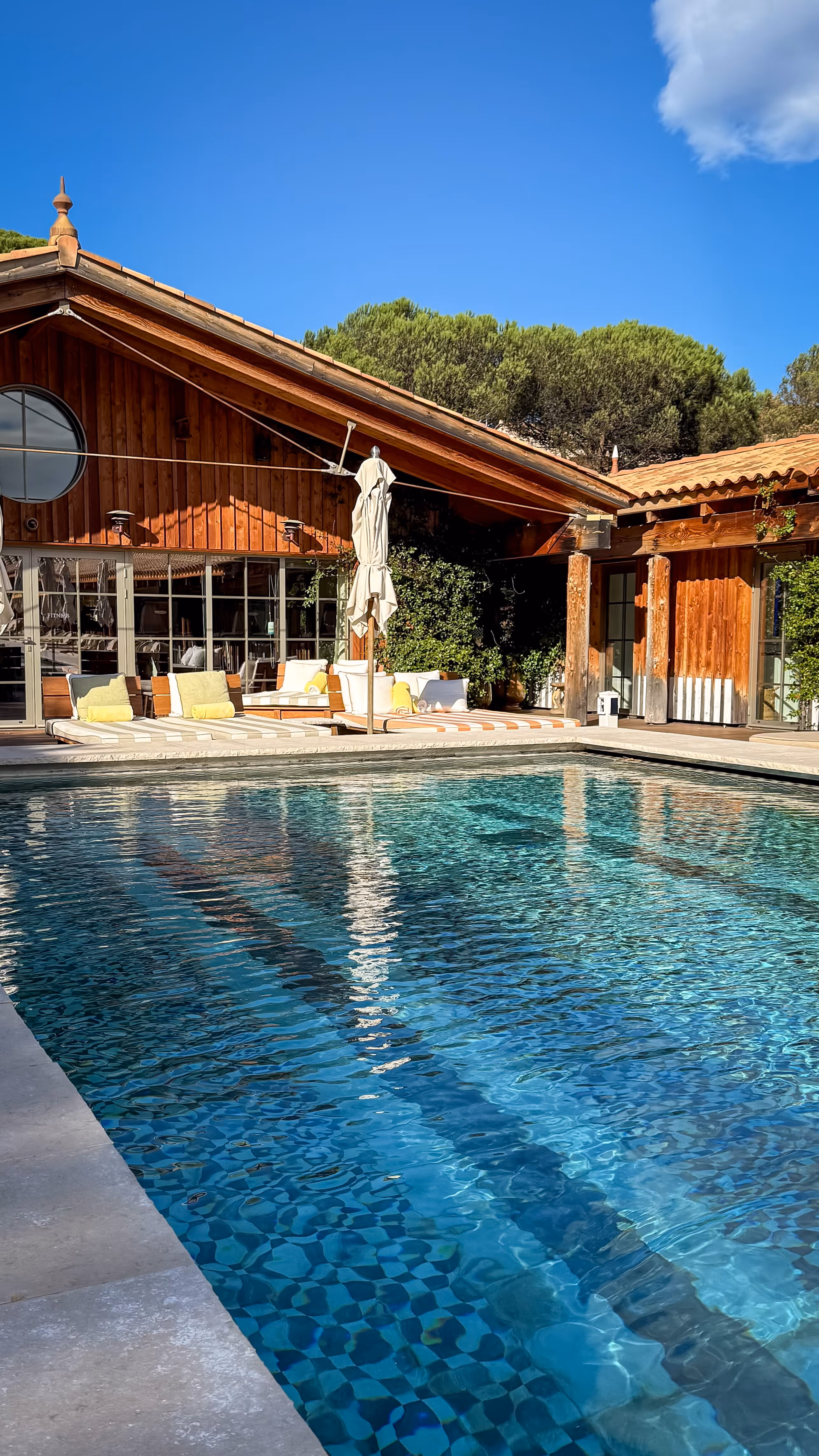 Clear blue swimming pool with checkered tiles beside a wooden cabin with outdoor lounge chairs and closed umbrellas.