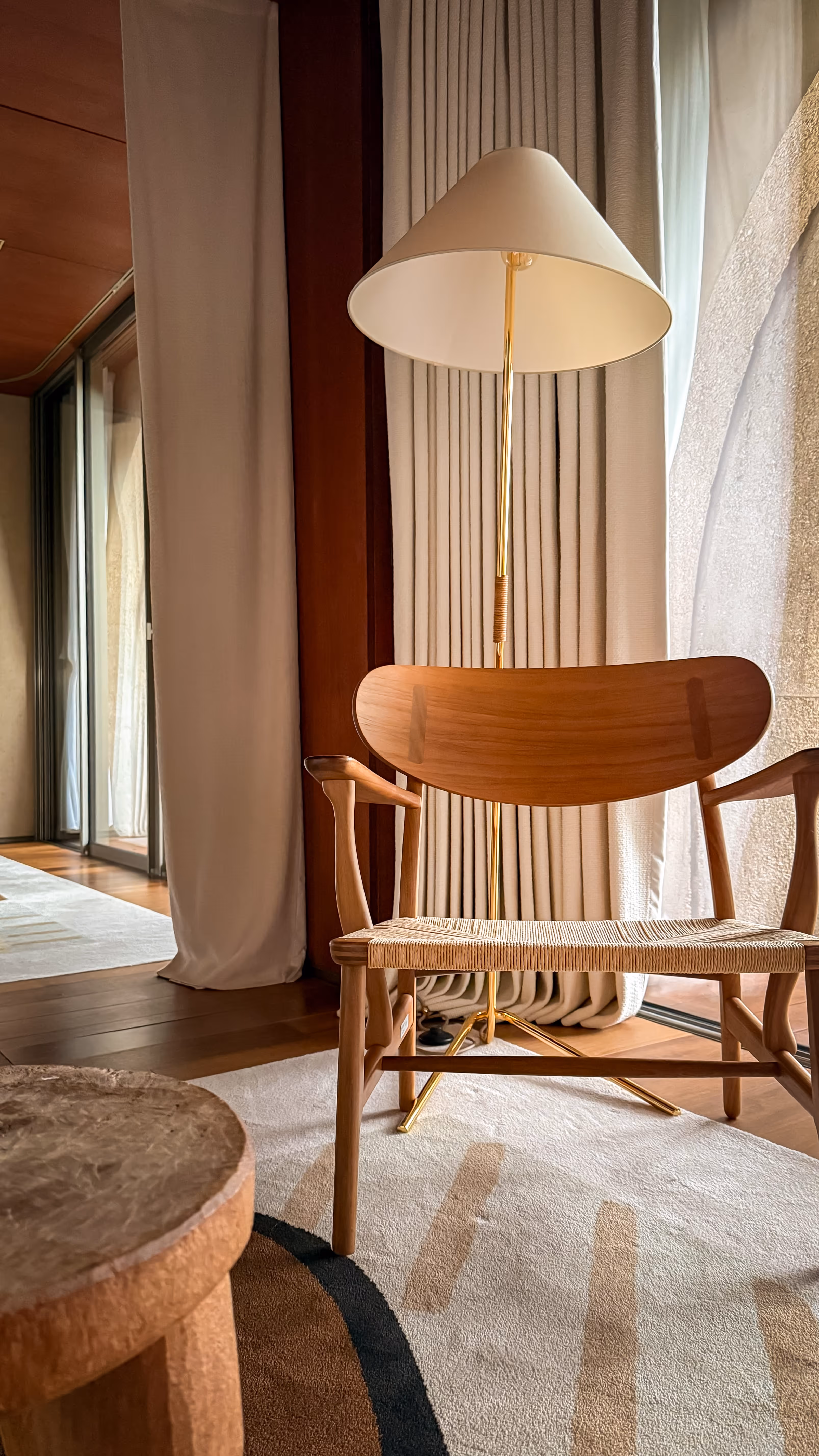 Wooden chair with woven seat in front of a beige floor lamp and curtains, standing on a patterned rug near a wooden side table.