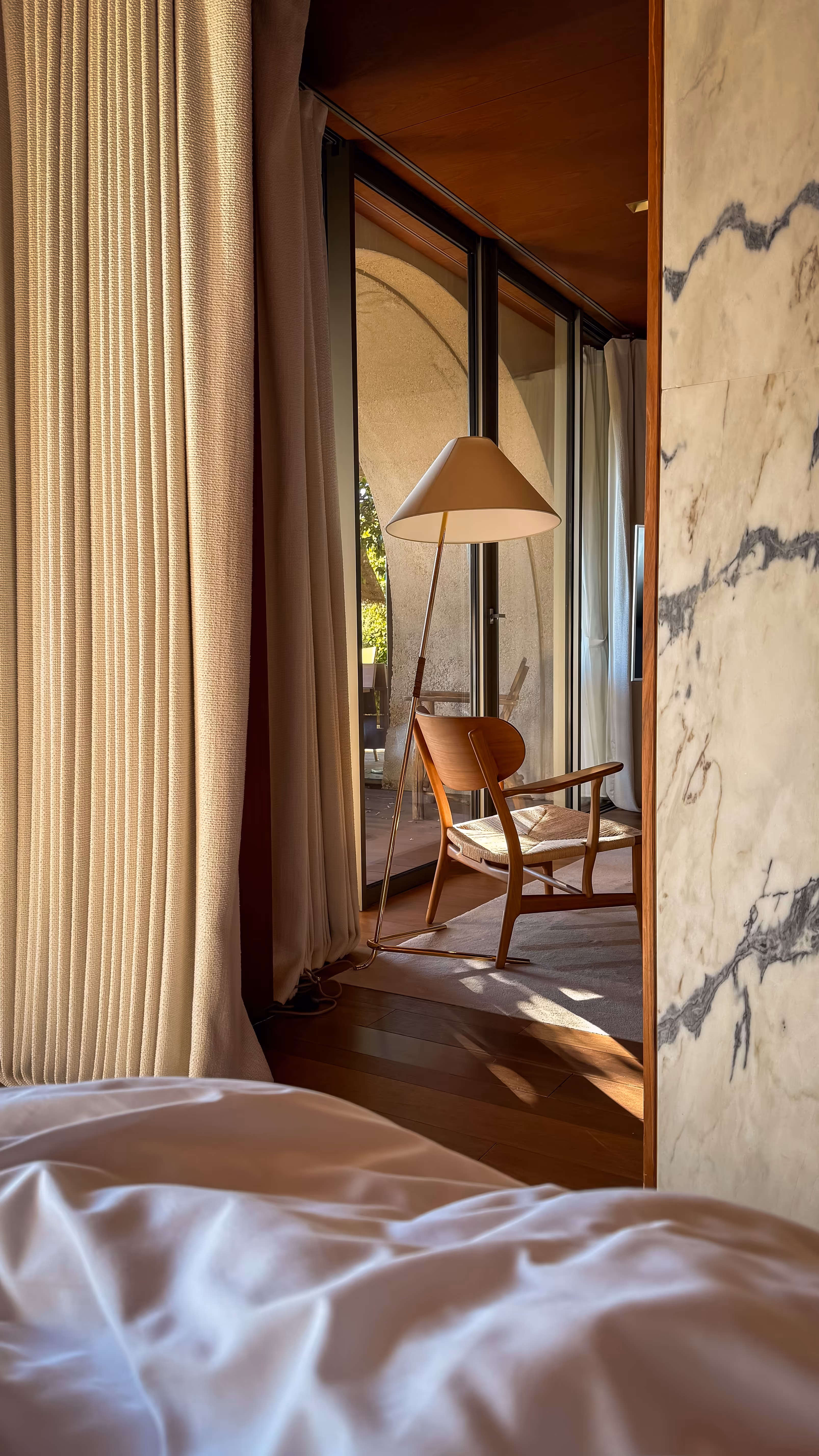 Modern wooden chair with woven seat next to a tall floor lamp by a glass door with curtains and a marble wall.