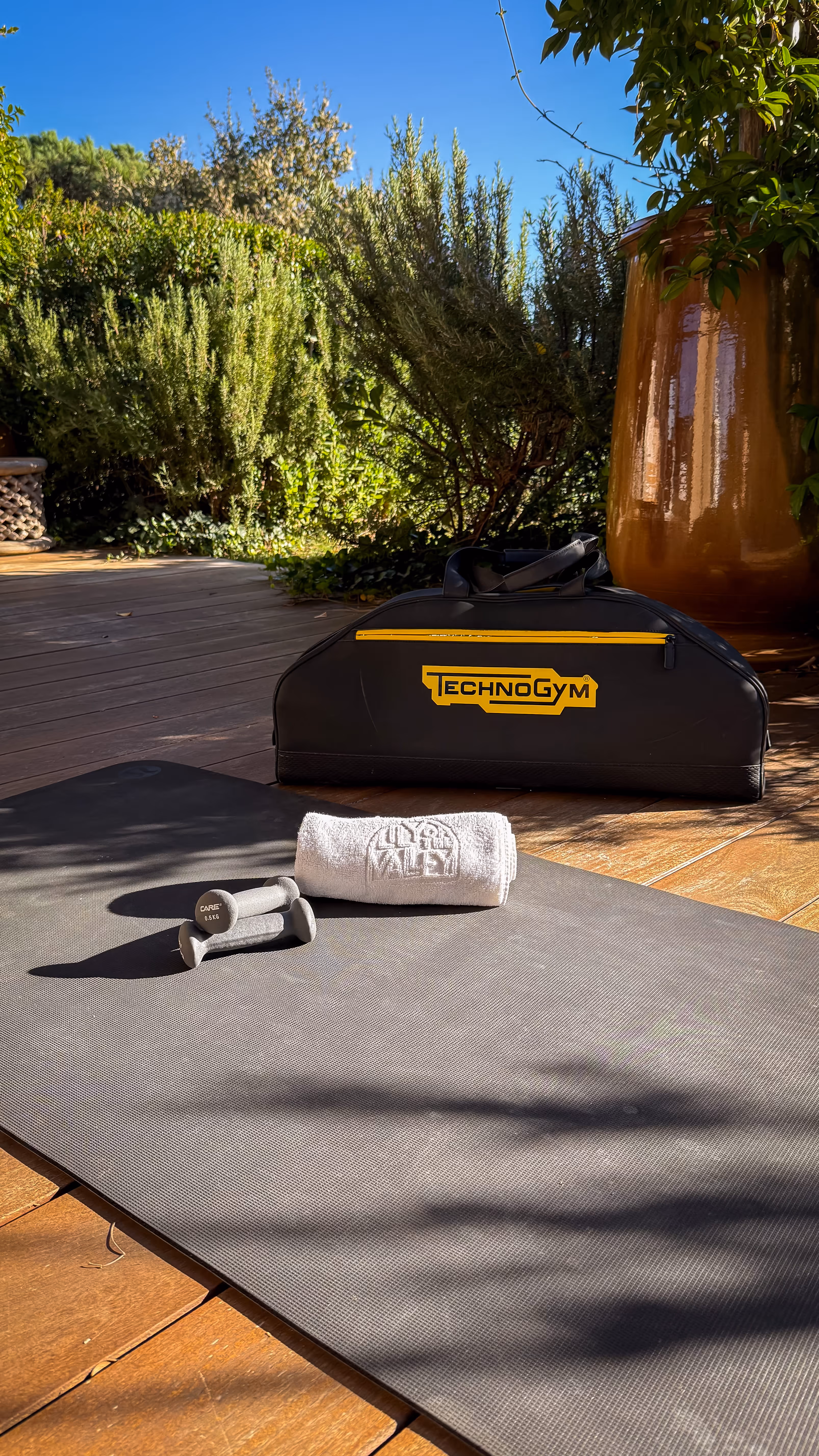 Outdoor workout setup with a black Technogym bag, a small gray dumbbell set, a rolled white towel, and a black exercise mat on wooden decking.
