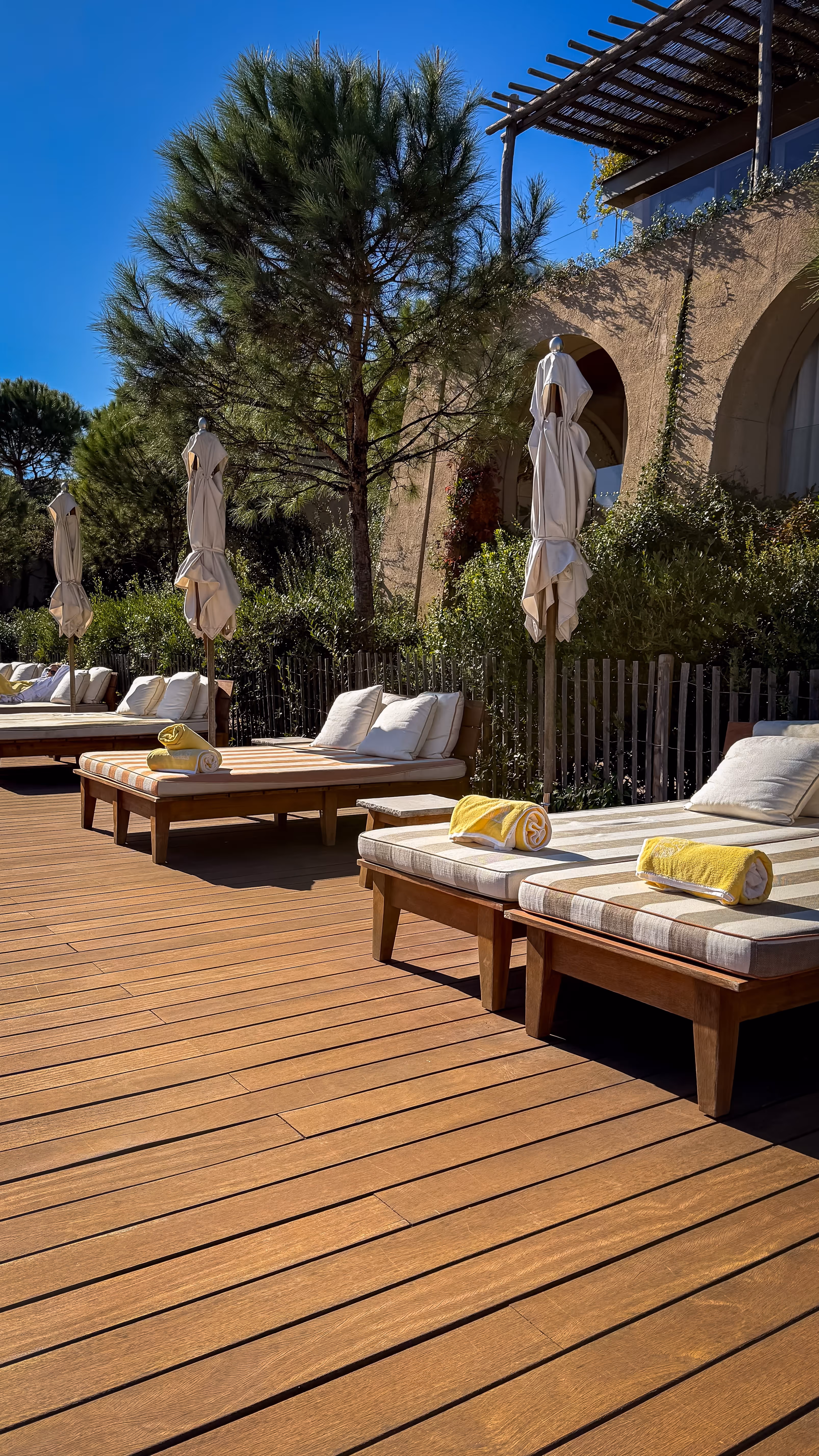 Sunlit outdoor lounge area with wooden deck, cushioned sunbeds with rolled yellow towels, closed umbrellas, and green trees.