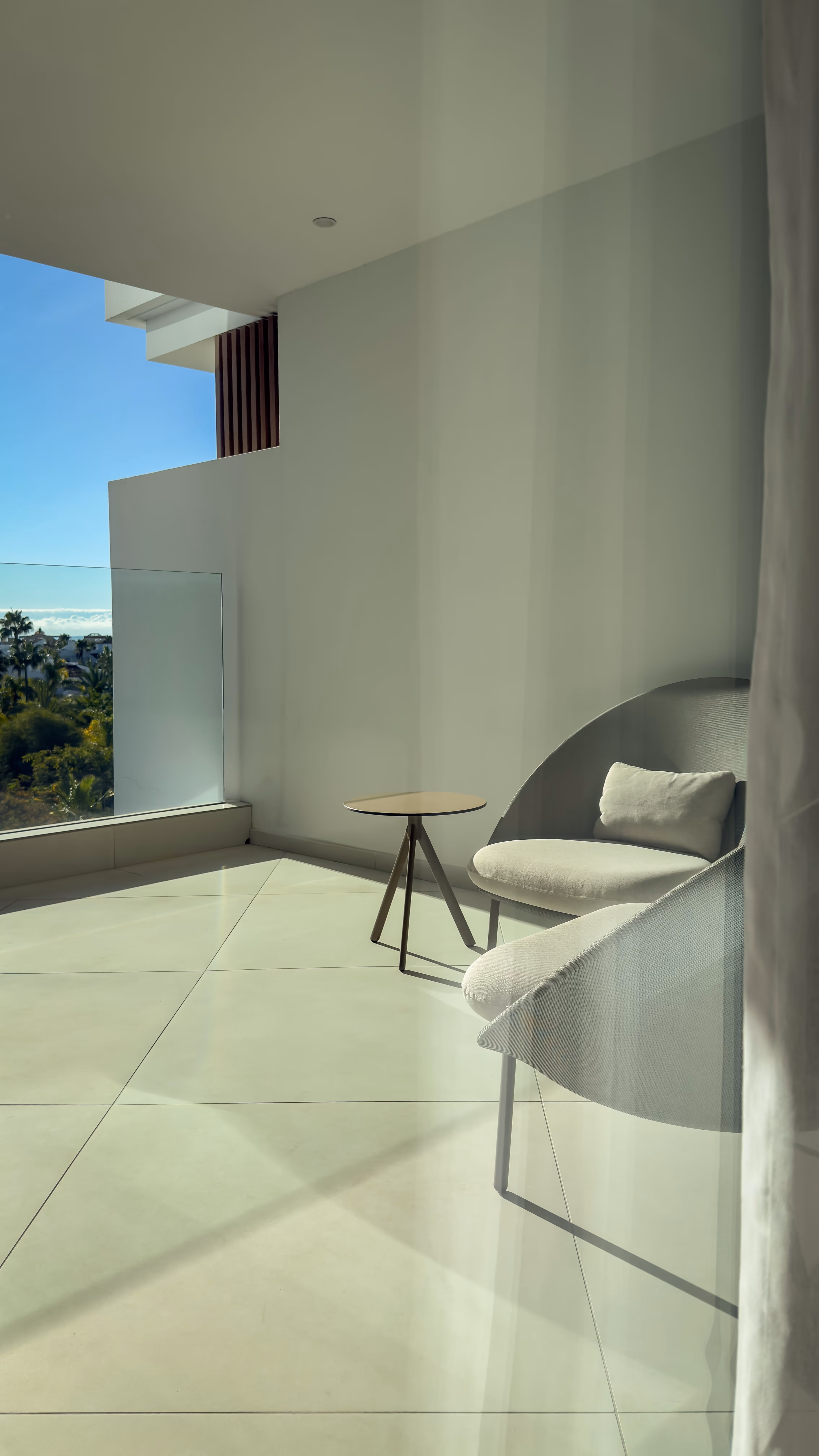 Minimalist balcony with beige tile floor, two cushioned chairs, and a small round wooden table, overlooking greenery and clear sky.