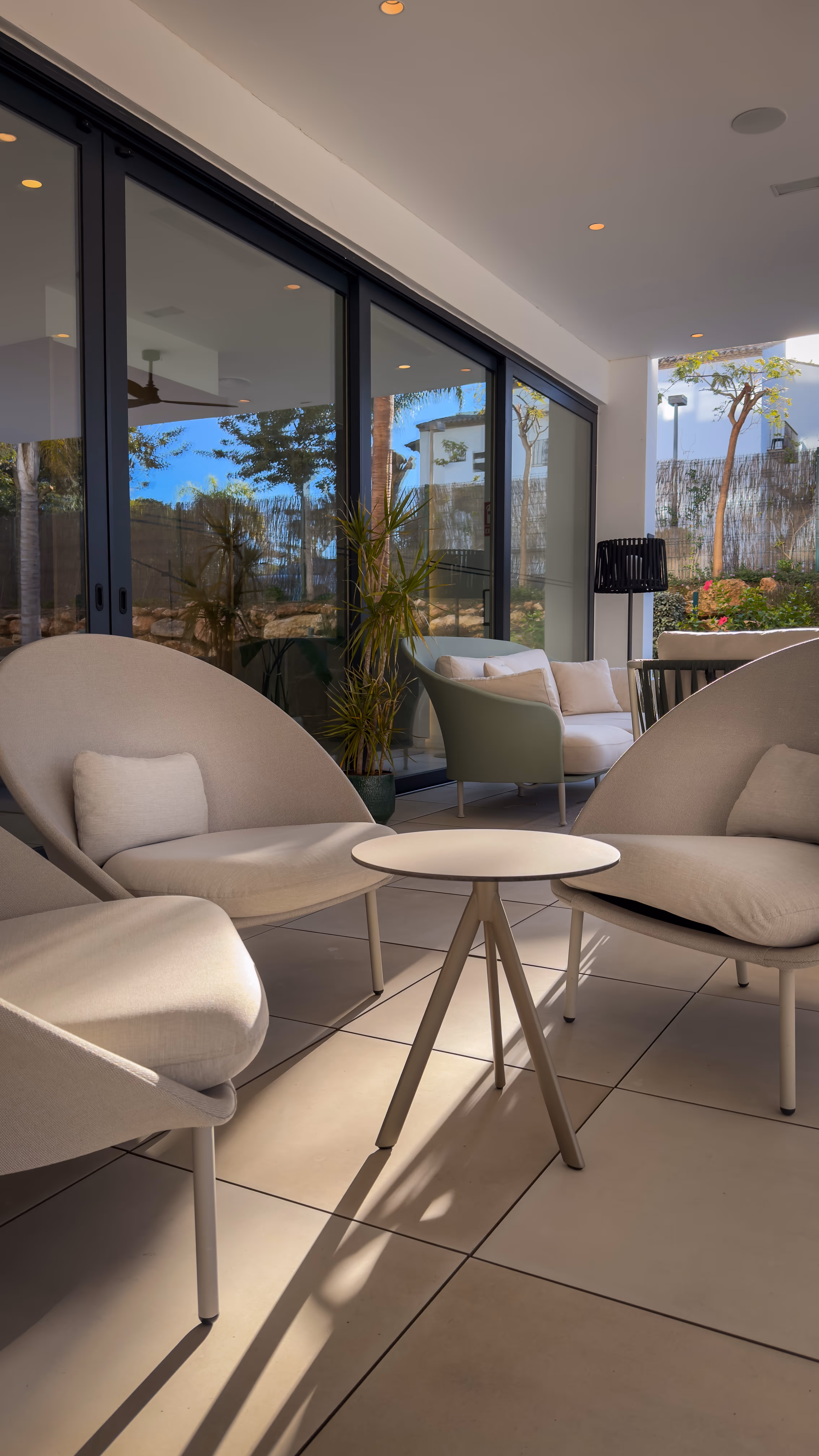 Outdoor patio area with beige cushioned lounge chairs around a small round table and glass sliding doors reflecting trees.