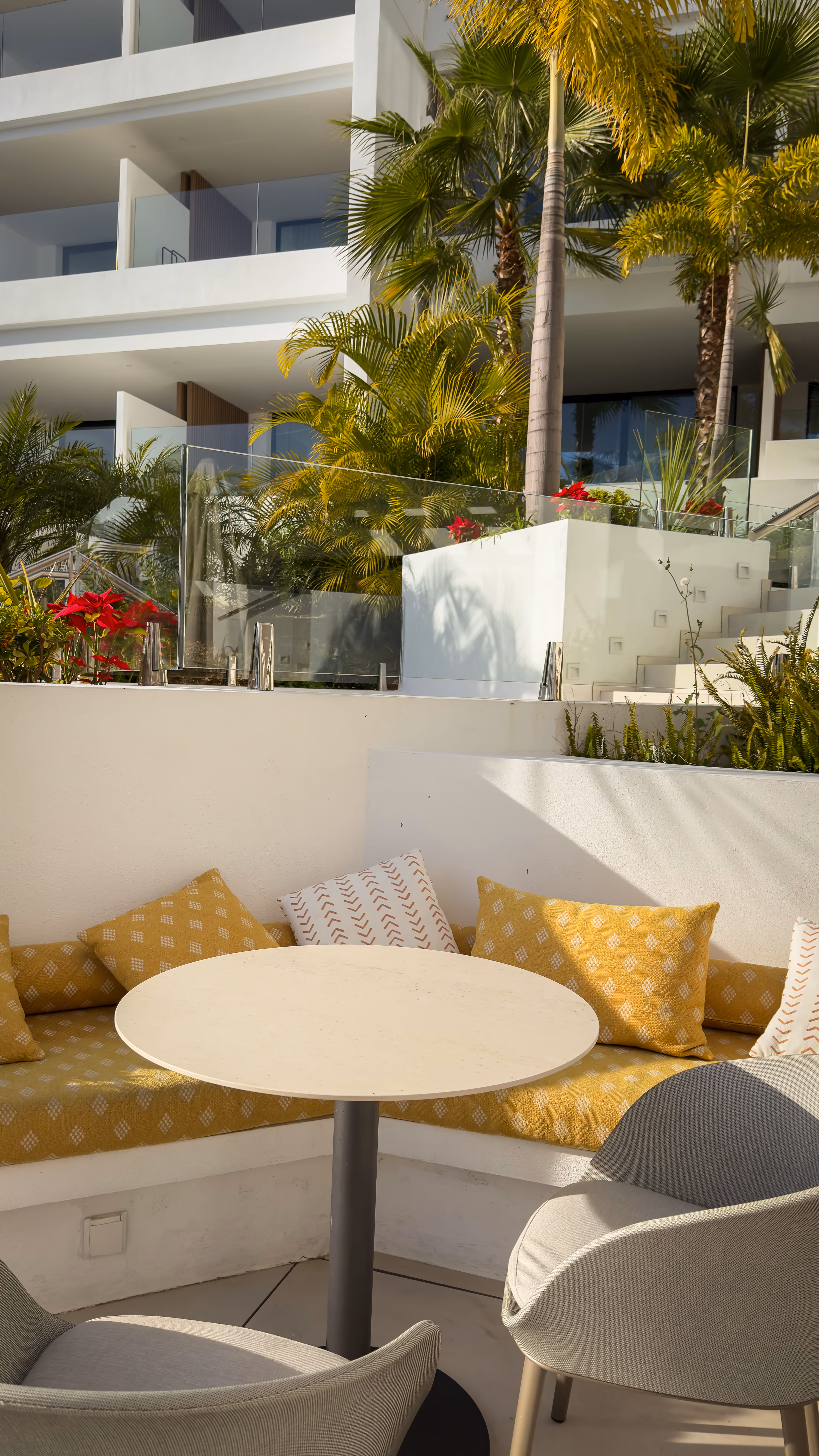 Outdoor seating area with a round white table, gray chairs, and a yellow cushioned bench with patterned pillows surrounded by tropical plants.